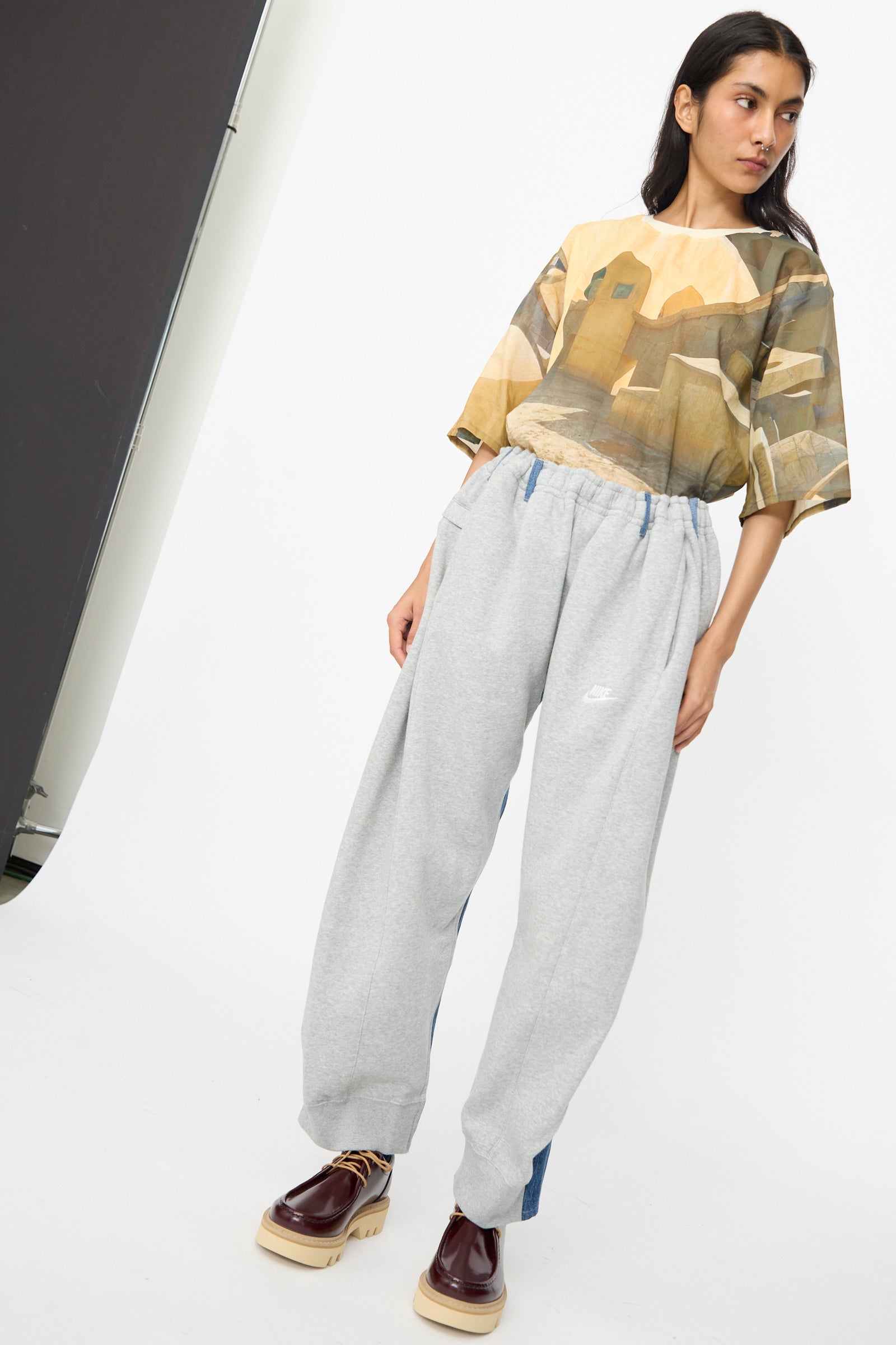 A woman stands against a plain backdrop wearing a printed t-shirt, No. 61 Overjoggingjeans in Lightgrey and Denimblue by Bless, and burgundy platform shoes.