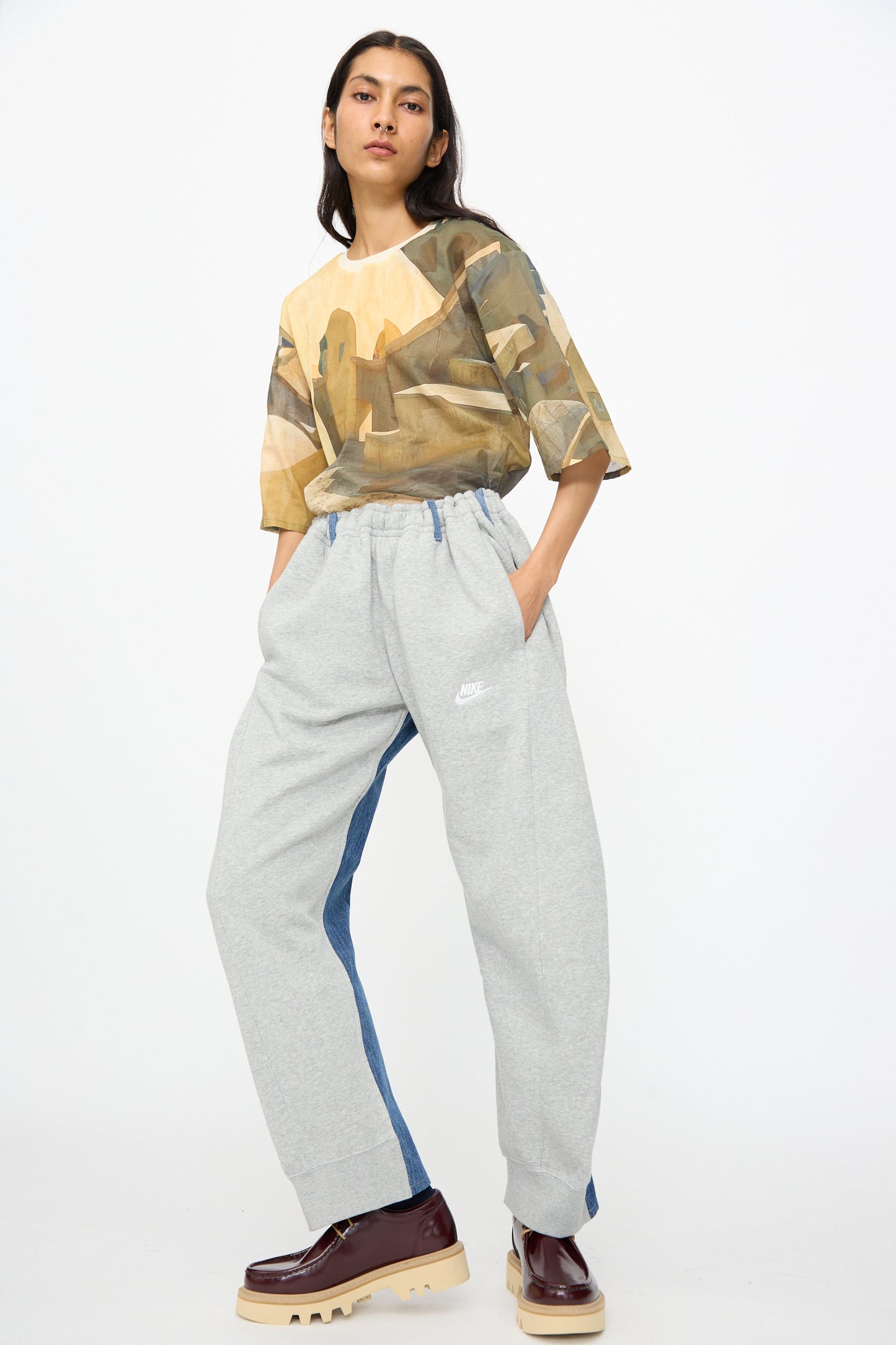 A person stands against a white background wearing a camouflage t-shirt, Bless No. 61 Overjoggingjeans in light grey and denim blue, and brown platform shoes.