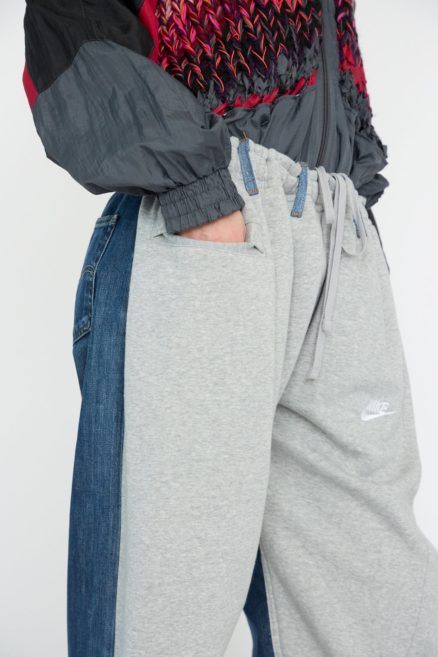 A model is wearing No. 61 Overjoggingjeans by Bless, featuring a blend of light grey and denim blue with a textured, multi-colored jacket and hands in their pockets.