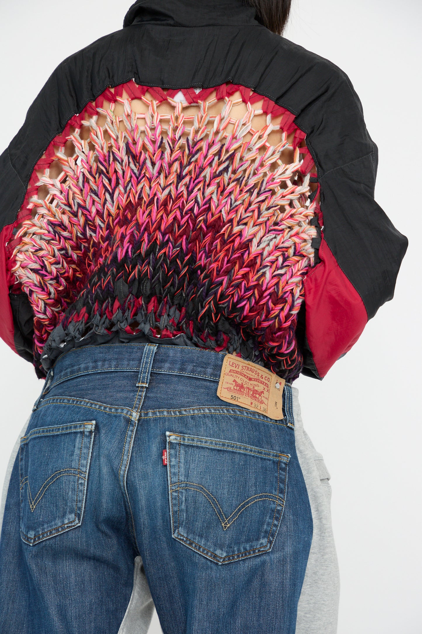 A model faces away wearing a black and red woven jacket with a multicolored back design, paired with Bless No. 61 Overjoggingjeans in Denimblue.