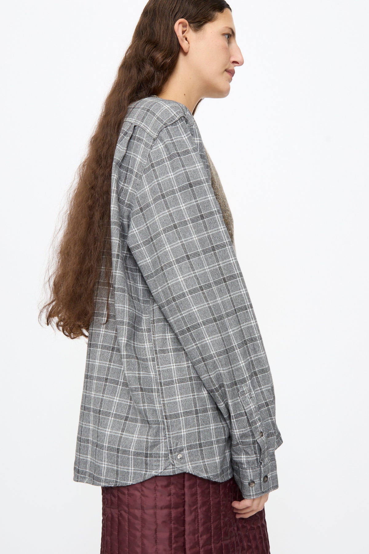Side profile of a person in a Bless No. 68 Frontinsertpullover in Unique I, a relaxed fit grey plaid flannel with a brown panel insert, layered over a quilted burgundy skirt.