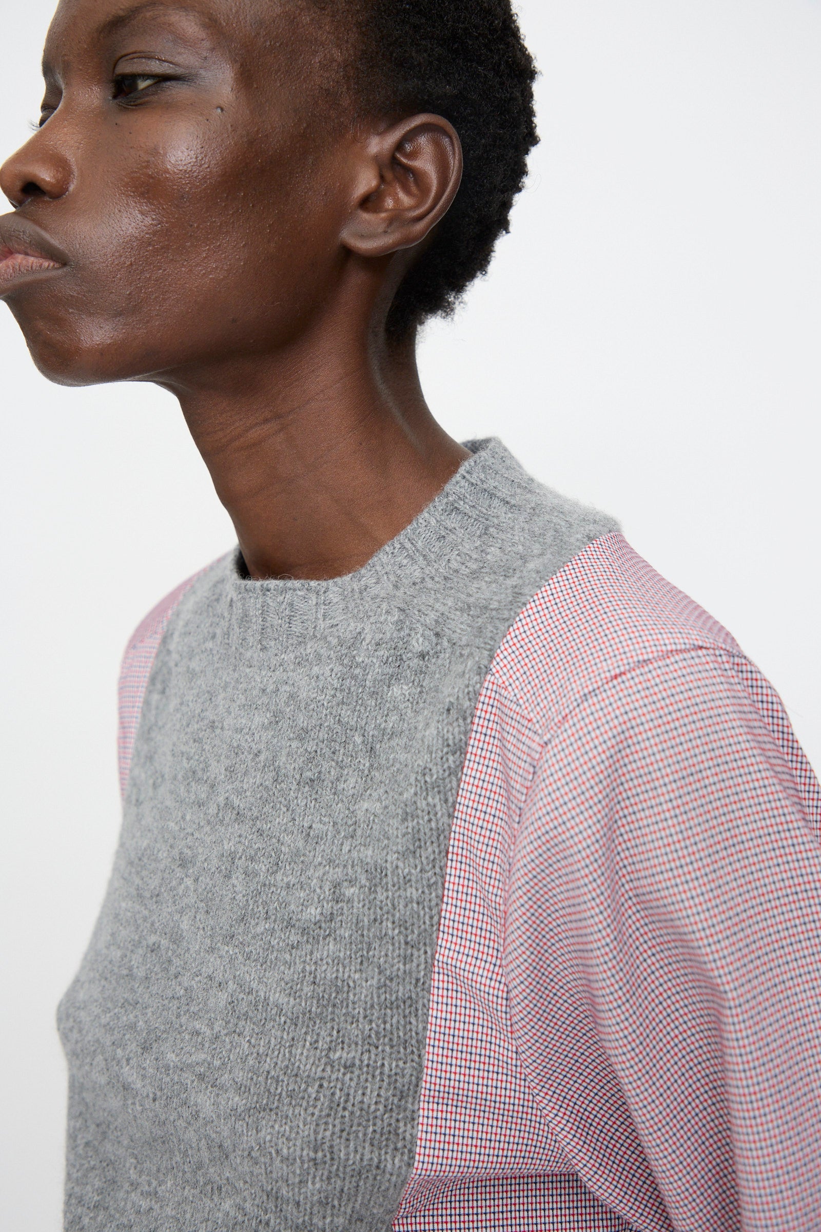 A person wears the Bless No. 68 Frontinsertpullover in Unique I, a grey shetland wool panel sleeveless sweater, layered over a pink patterned cotton poplin shirt, shown from the side against a plain background.