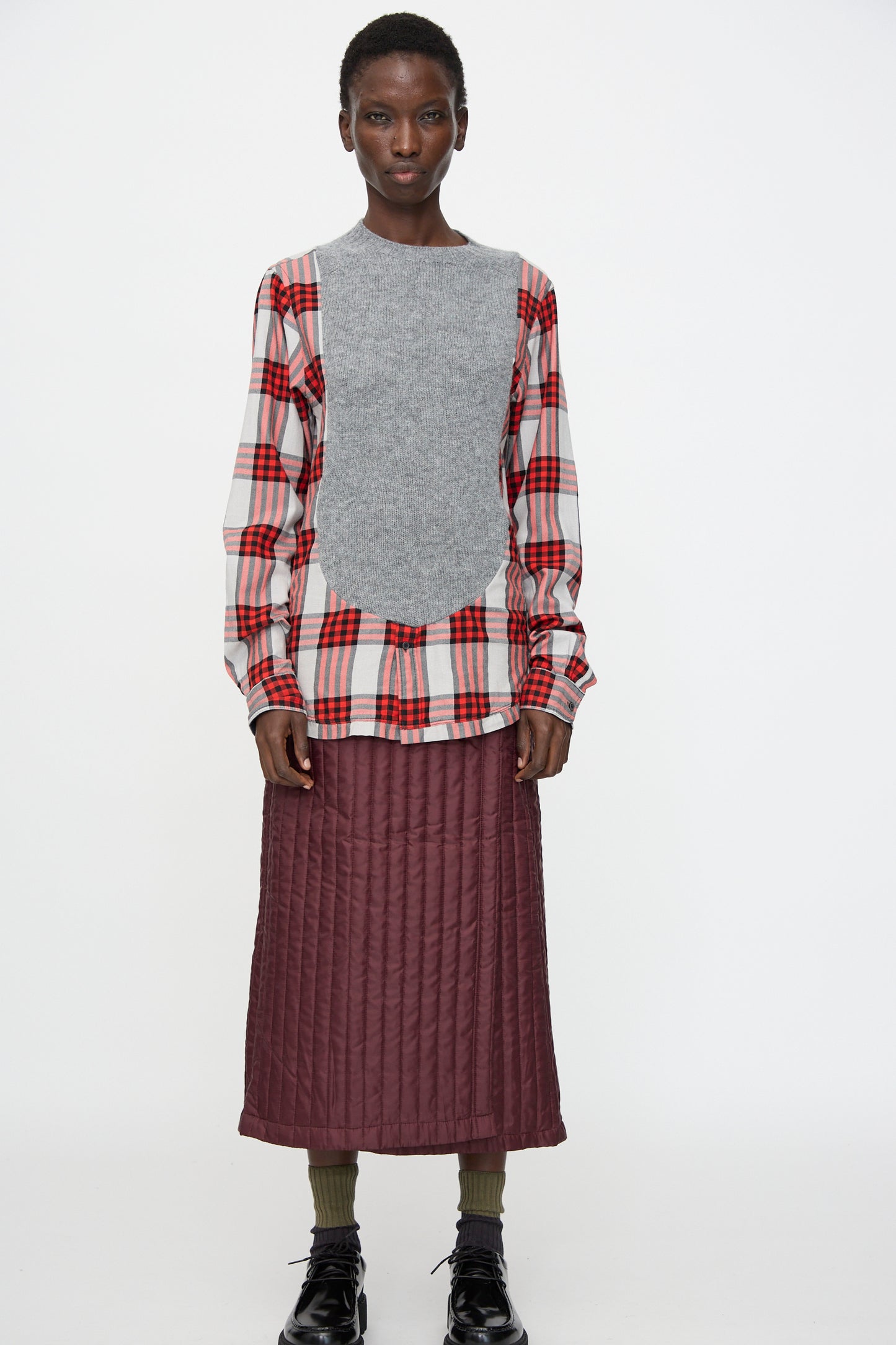 A person stands facing forward wearing the Bless No. 68 Frontinsertpullover in Unique II over a relaxed fit red flannel shirt, a maroon quilted skirt, green socks, and black shoes against a white background.