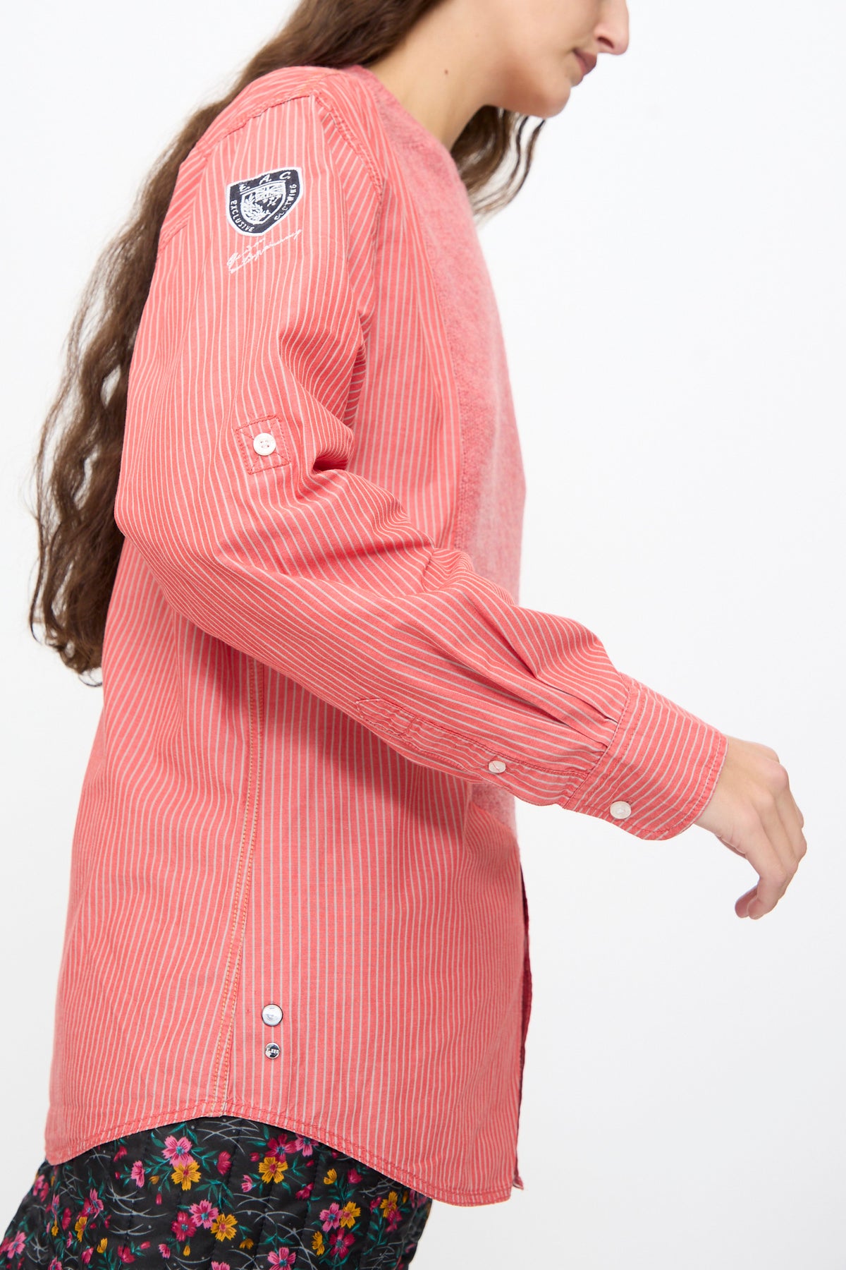 Side view of a person wearing a Bless No. 68 Frontinsertpullover in Unique II, a pink, white-striped poplin pullover with a textured front panel, a circular shoulder emblem, and button-down detailing.