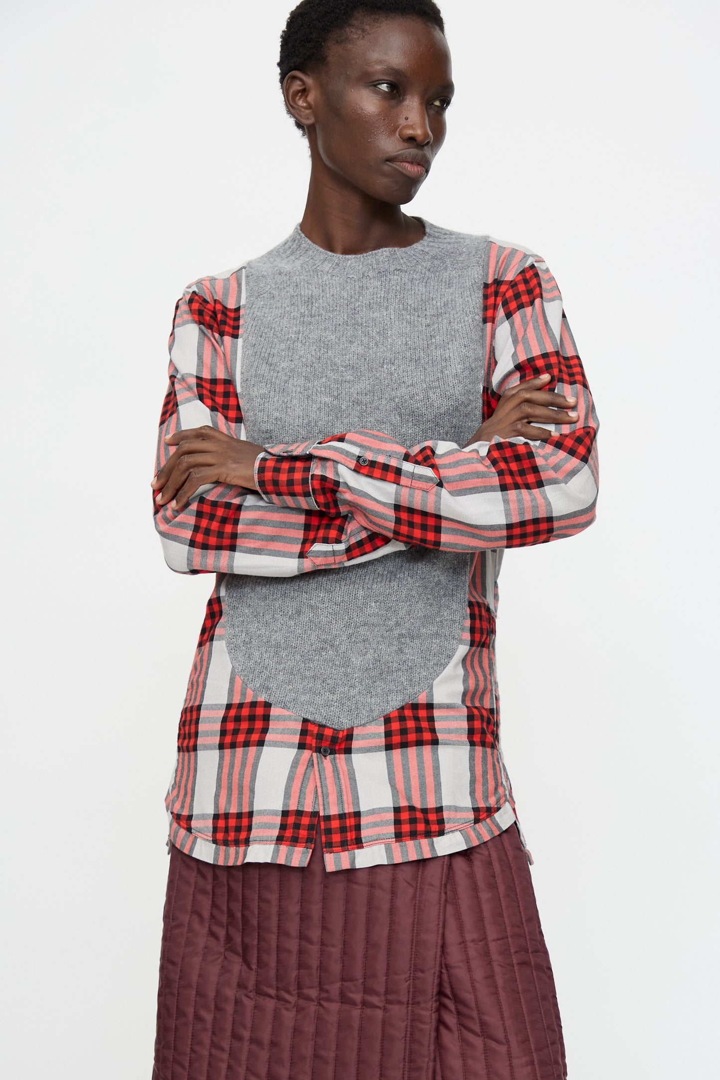 A person stands with arms crossed wearing the Bless No. 68 Frontinsertpullover in Unique II and a maroon quilted skirt, against a plain white background.