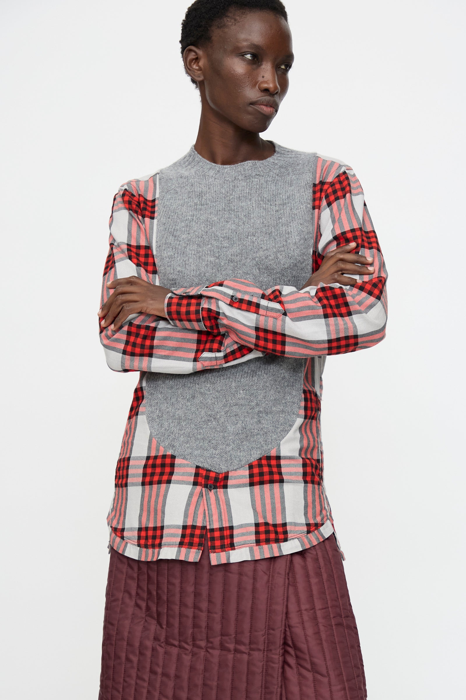 A person stands with arms crossed wearing the Bless No. 68 Frontinsertpullover in Unique II and a maroon quilted skirt, against a plain white background.