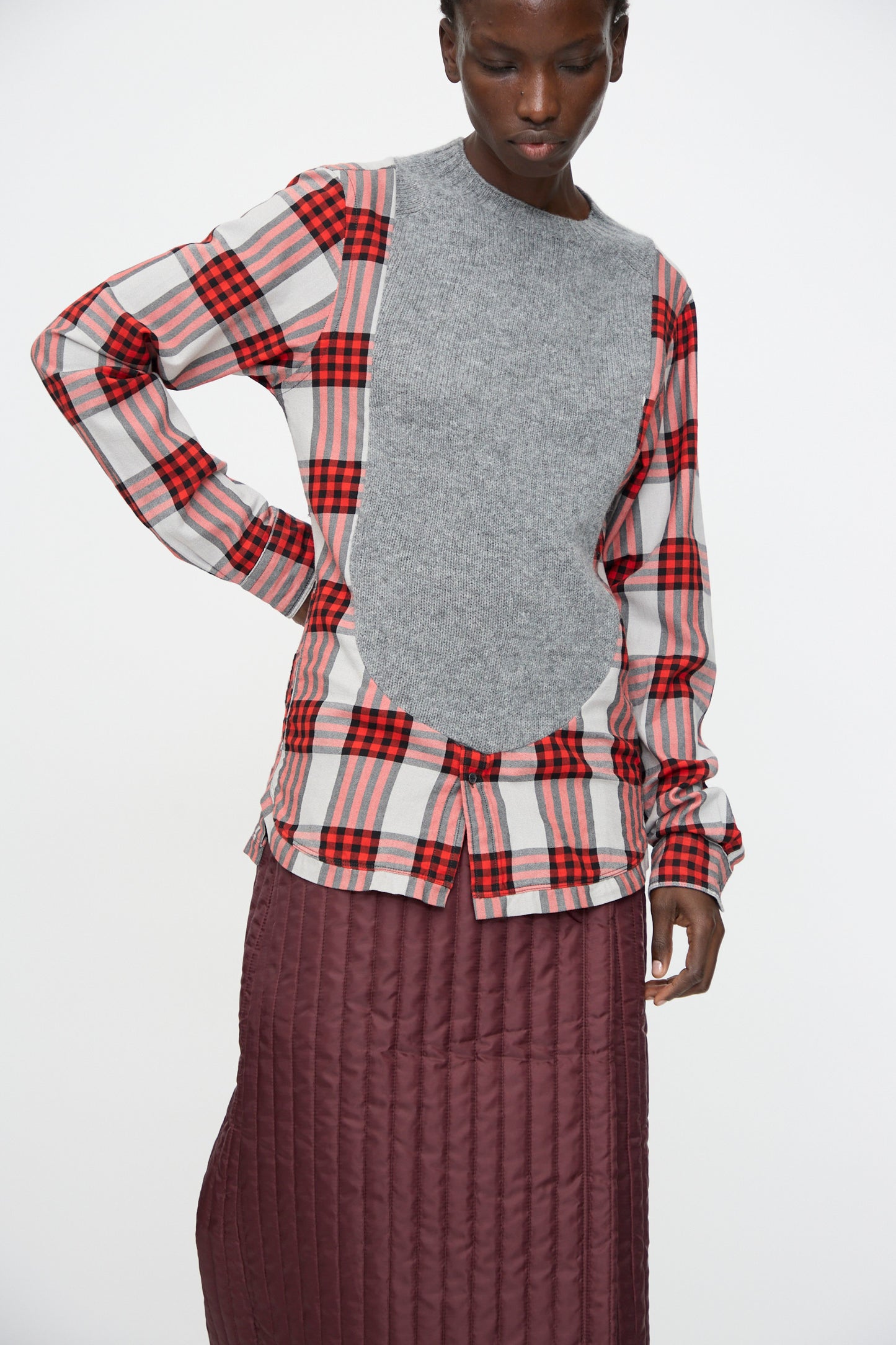 A person stands against a plain white background wearing the Bless No. 68 Frontinsertpullover in Unique II, a relaxed fit red and grey plaid flannel shirt with a grey knit front panel, paired with a maroon quilted skirt.