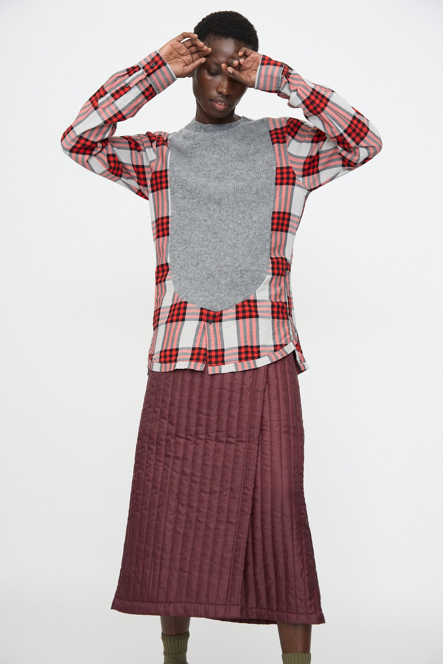 A person wears the Bless No. 68 Frontinsertpullover in Unique II—a gray and red plaid flannel shirt with a gray panel—paired with a burgundy quilted skirt and green socks, standing against a white background.