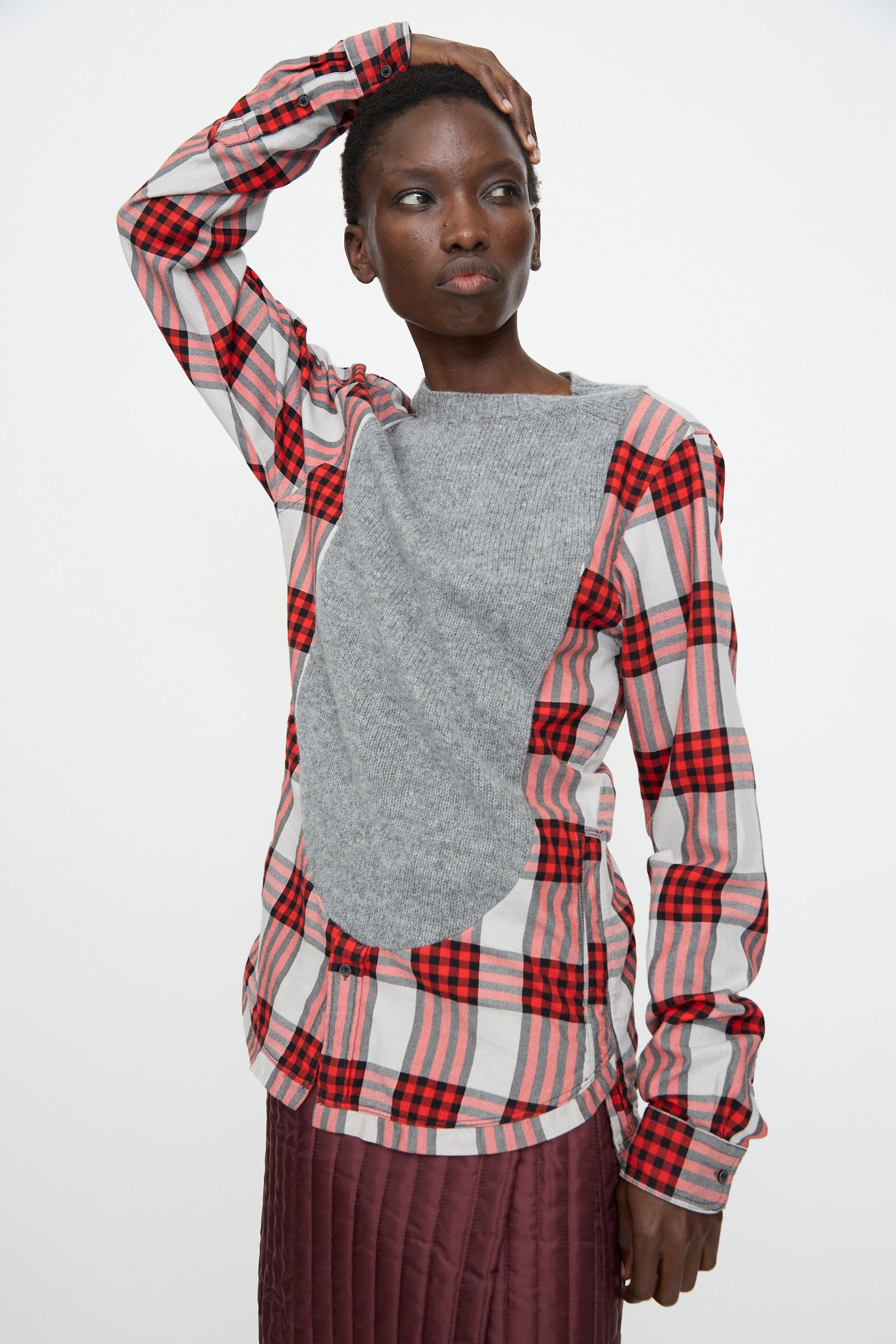 A person wearing the Bless No. 68 Frontinsertpullover in Unique II, a relaxed fit red plaid flannel shirt with a large gray Shetland wool panel on the front, paired with a maroon quilted skirt, stands with one hand on their head against a white background.
