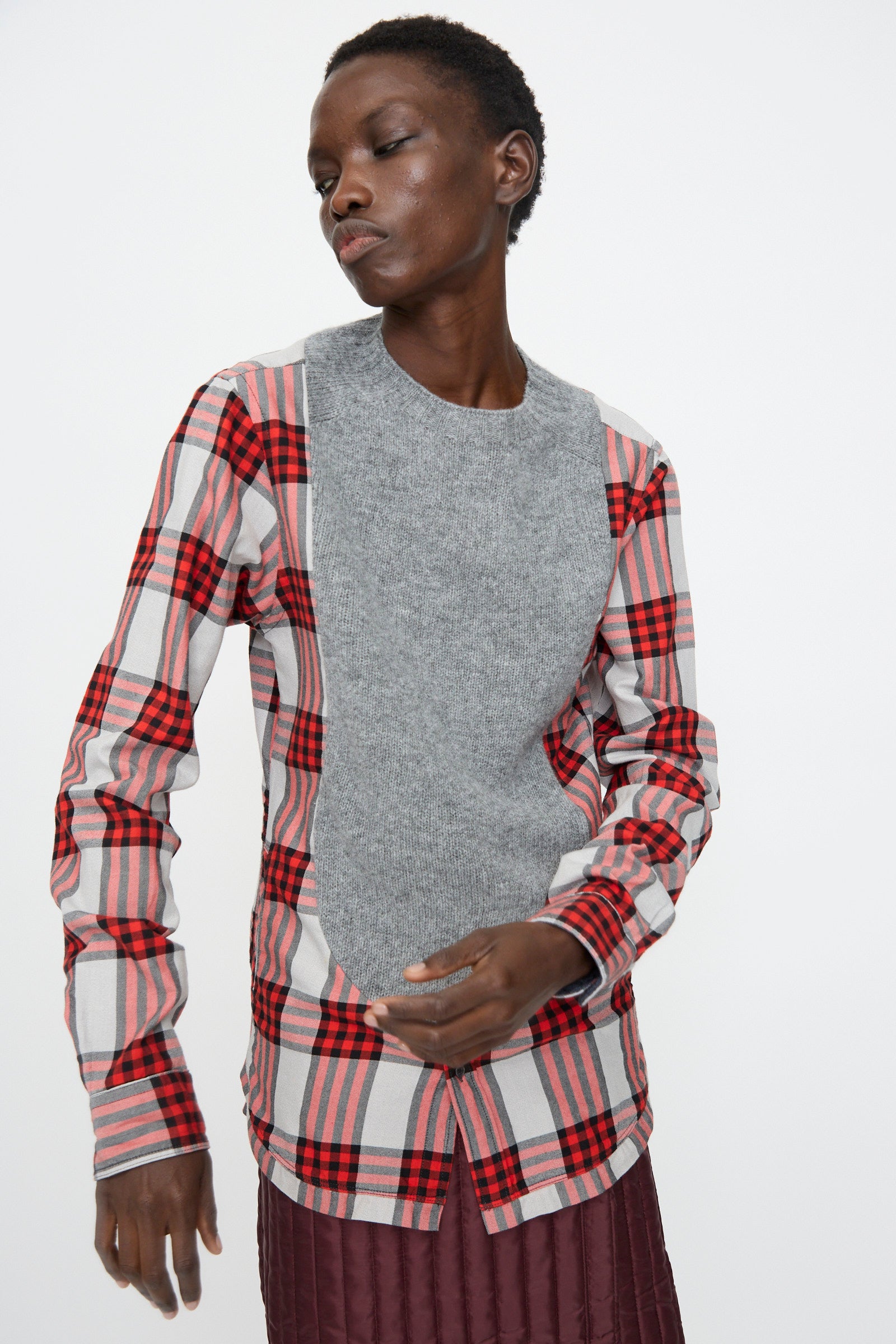 A person wears the Bless No. 68 Frontinsertpullover in Unique II, featuring red and gray plaid sleeves and sides with a solid gray front panel, paired with a burgundy skirt, standing against a plain white background.