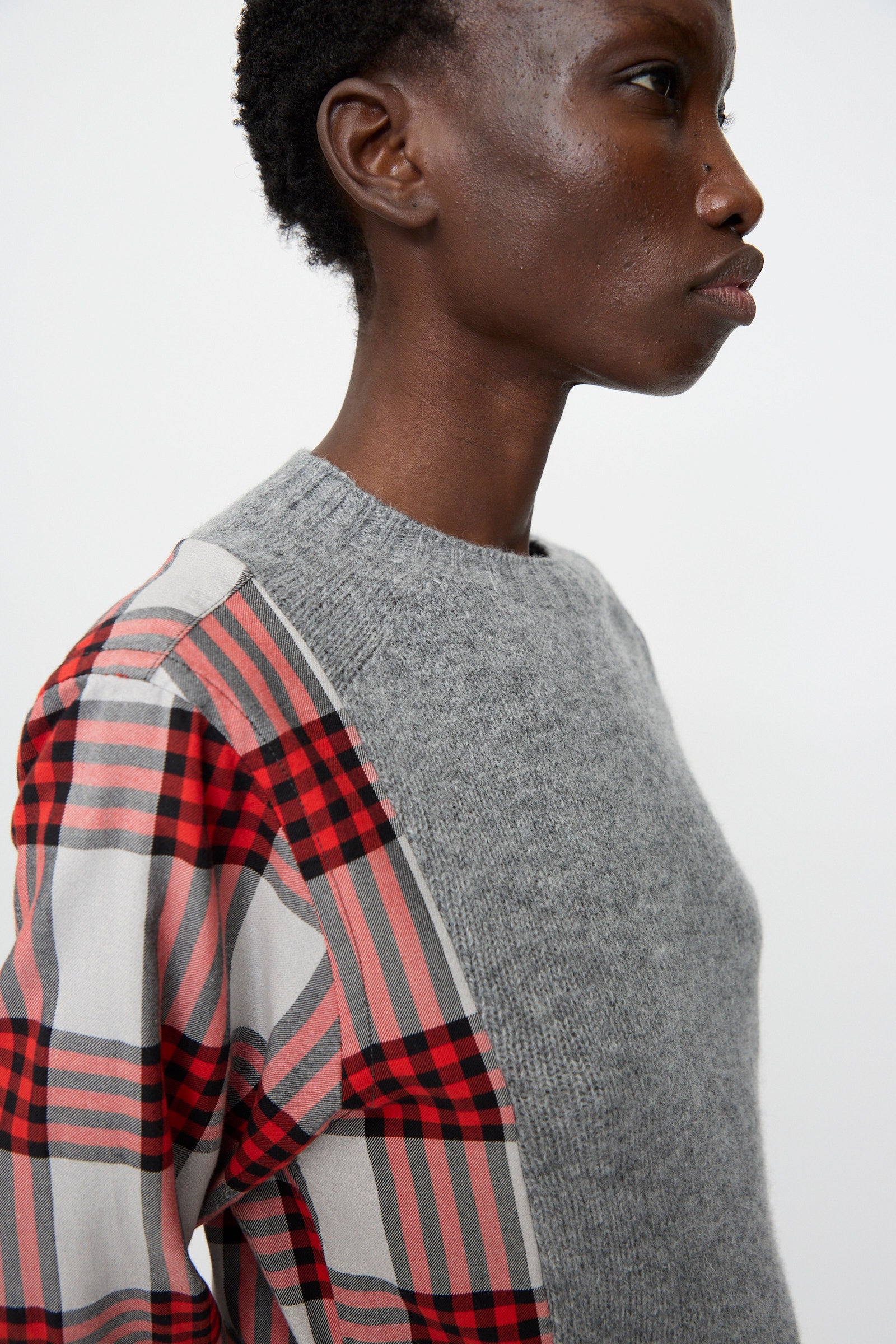 A person wears the Bless No. 68 Frontinsertpullover in Unique II—a gray Shetland wool knit sweater with one sleeve in red and black plaid flannel—shown from the side against a plain background, relaxed fit.