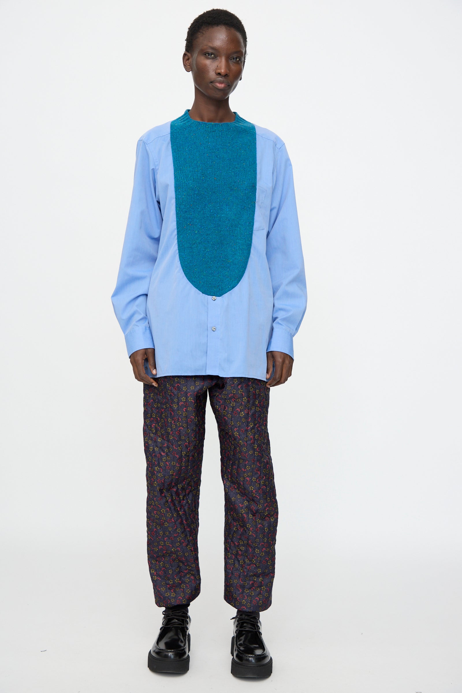A person stands against a white background wearing the Bless No. 68 Frontinsertpullover in Unique III, paired with patterned pants and black platform shoes.