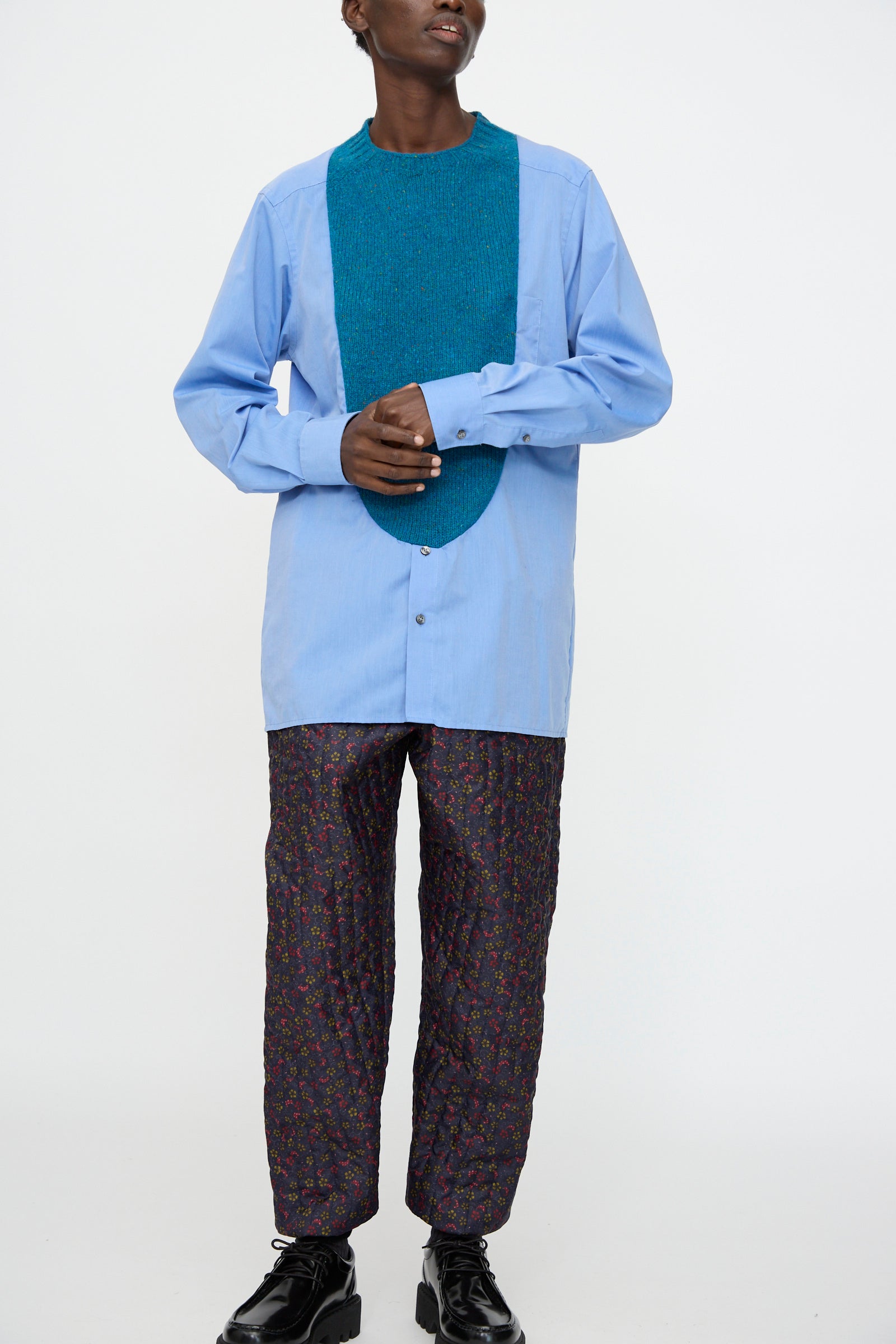 Person wearing Bless's No. 68 Frontinsertpullover in Unique III—a relaxed fit, light blue cotton poplin shirt with a large teal Shetland wool panel—styled with patterned quilted pants and black shoes, standing against a plain white background.