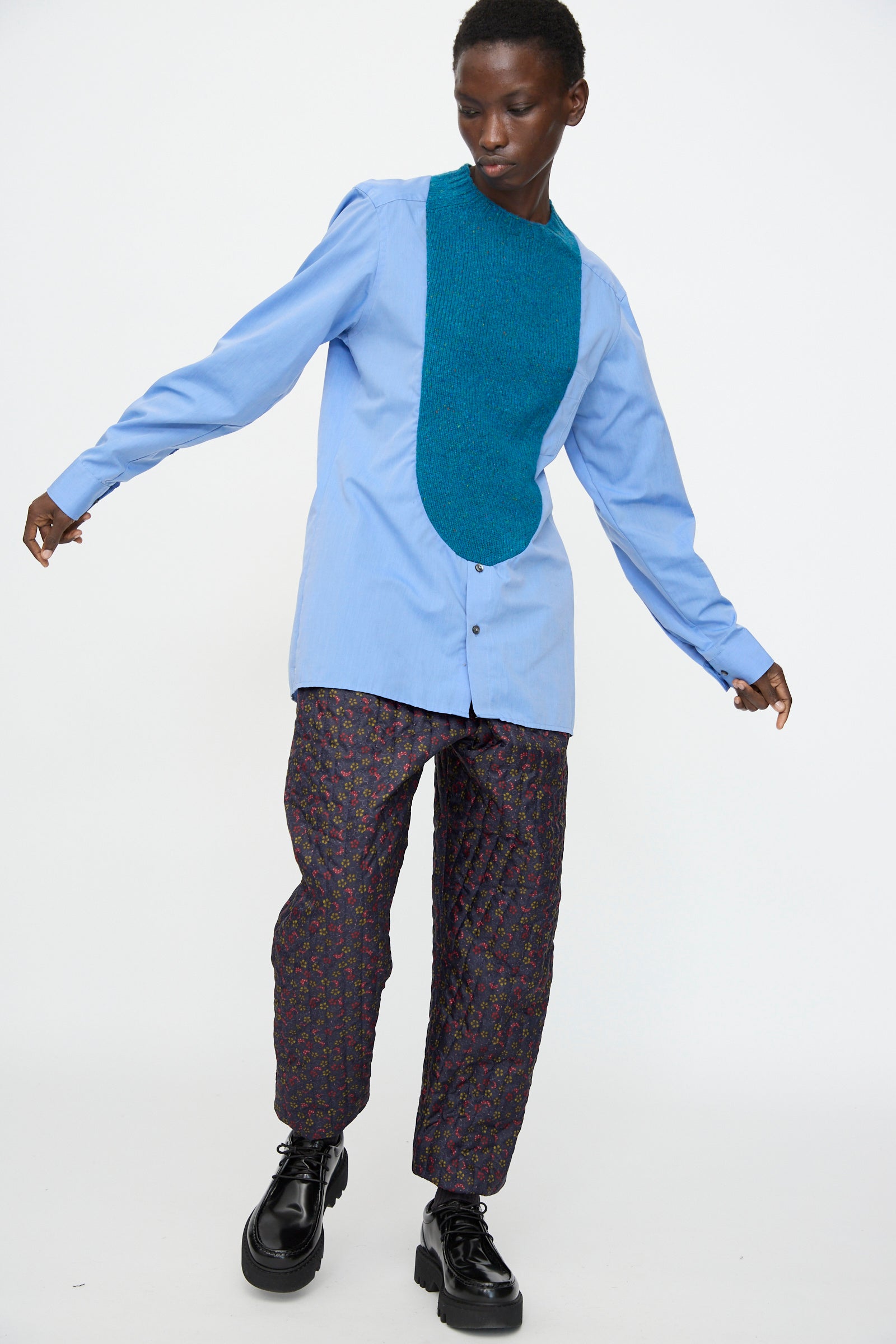A person poses against a plain background wearing the Bless No. 68 Frontinsertpullover in Unique III, styled with a teal scarf, patterned dark pants, black shoes, arms extended and one leg slightly raised.