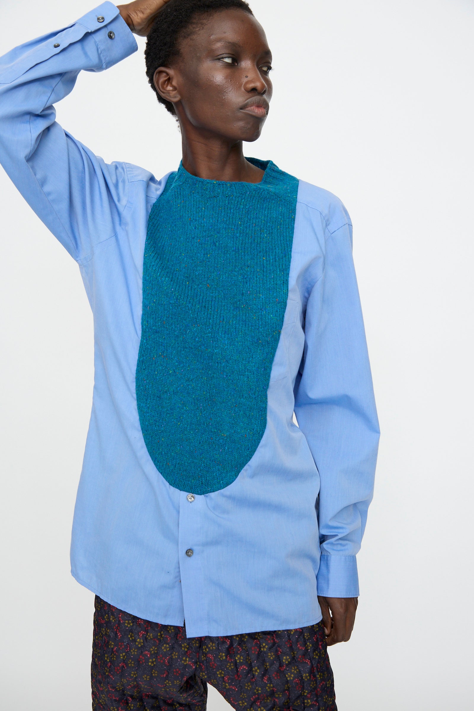 A person wears the Bless No. 68 Frontinsertpullover in Unique III, a relaxed fit light blue cotton poplin shirt with a textured Shetland wool front panel, paired with patterned dark pants, standing against a plain white background.