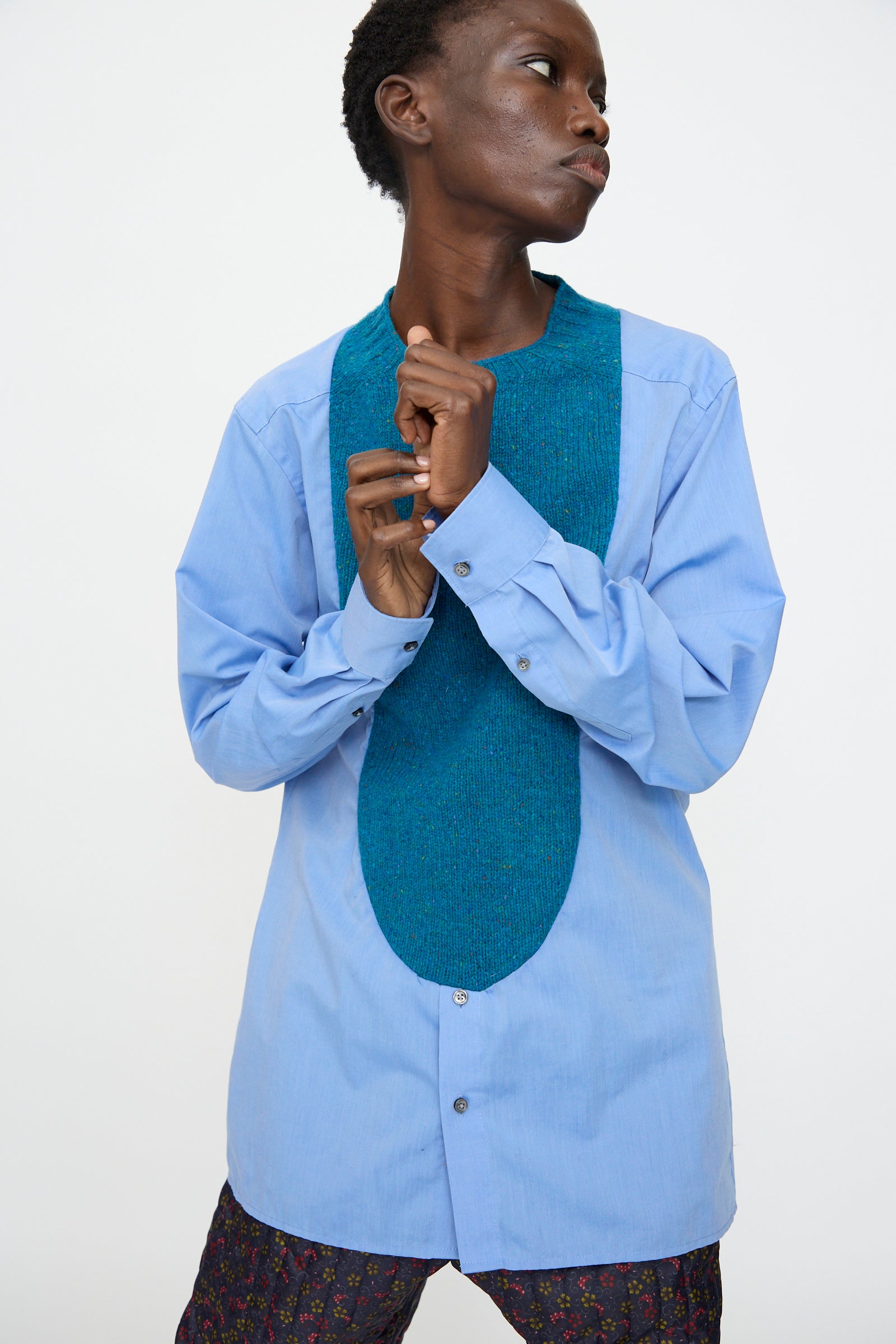A person wears the Bless No. 68 Frontinsertpullover in Unique III, a relaxed fit light blue shirt with a textured oval chest panel, paired with patterned pants against a plain white background.