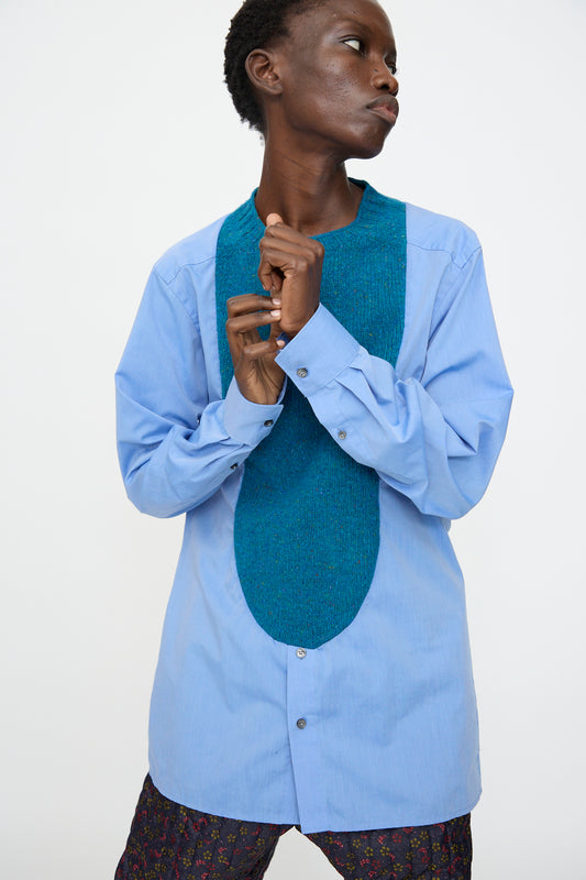 A person wears the Bless No. 68 Frontinsertpullover in Unique III, a relaxed fit light blue shirt with a textured oval chest panel, paired with patterned pants against a plain white background.