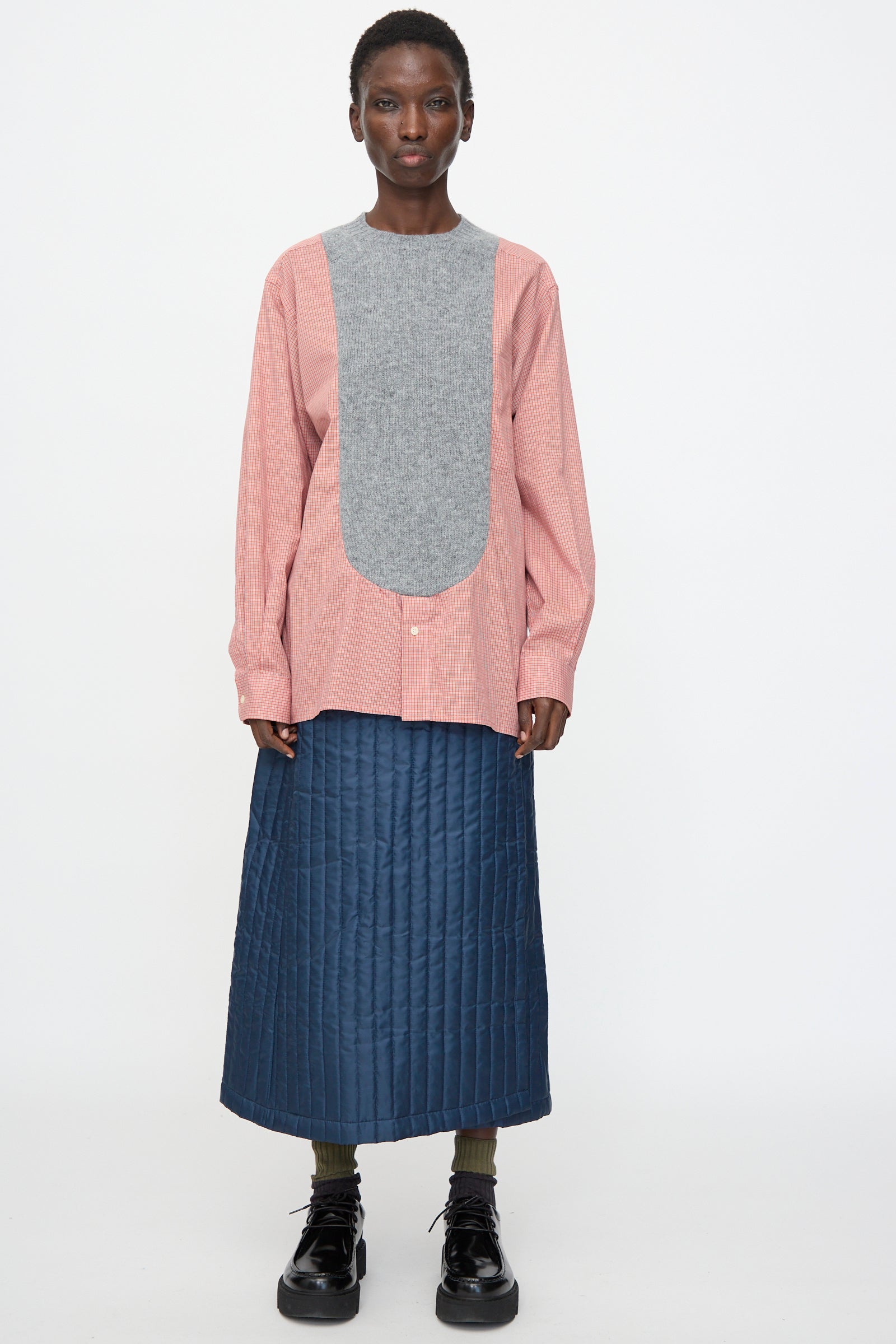 A person faces forward wearing the Bless No. 68 Frontinsertpullover in Unique IV with a gray bib, styled with a blue quilted skirt, green socks, and black shoes against a plain white background.