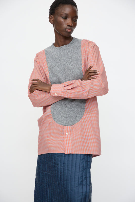 A person stands with arms crossed, wearing the Bless No. 68 Frontinsertpullover in Unique IV—a pink checkered cotton poplin shirt with a large gray wool panel—paired with a blue quilted skirt, set against a plain white background.
