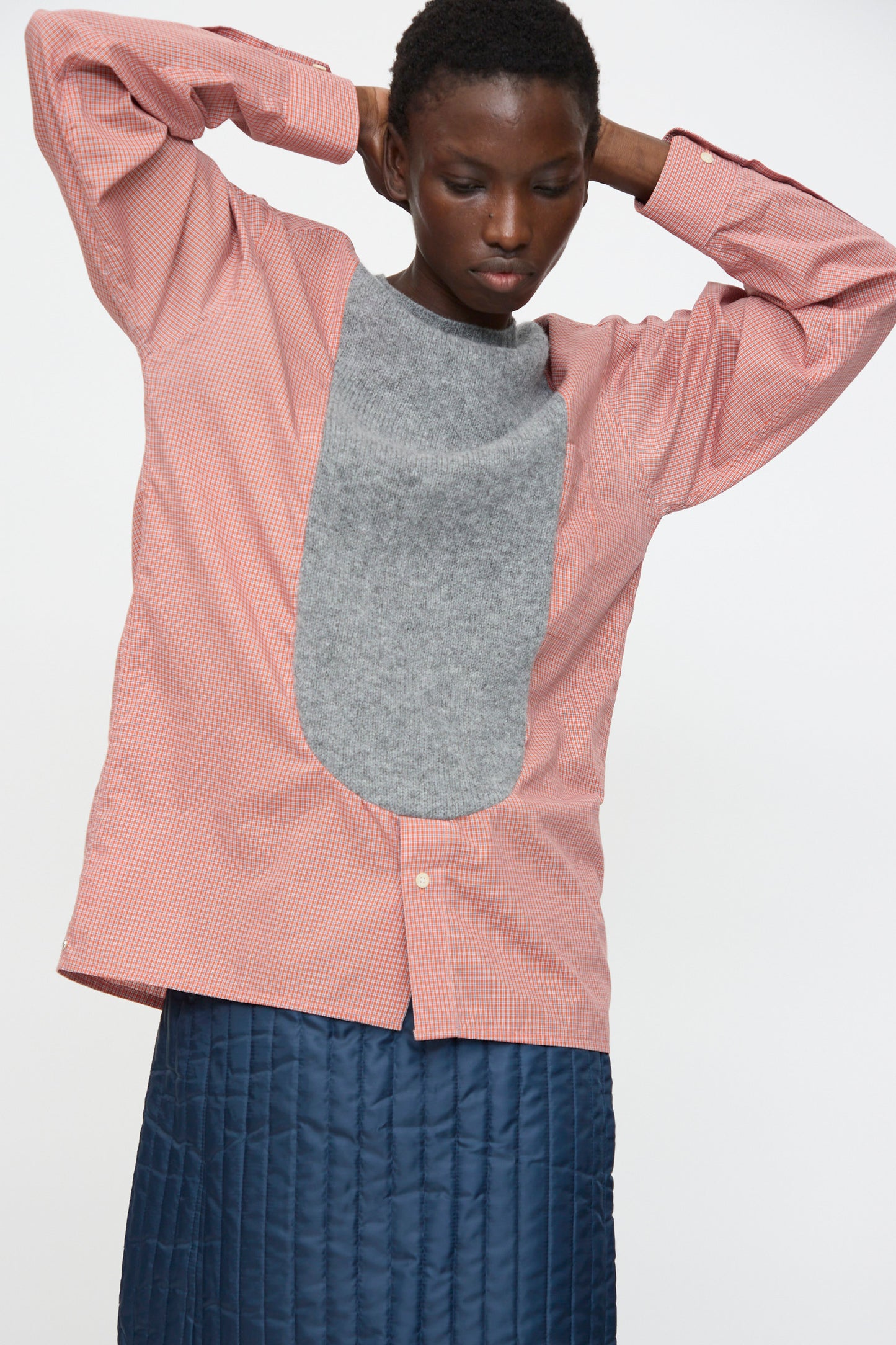 A person wears the Bless No. 68 Frontinsertpullover in Unique IV, featuring a red and white cotton poplin shirt with a large Shetland wool front panel, paired with a blue quilted skirt, standing against a plain background.