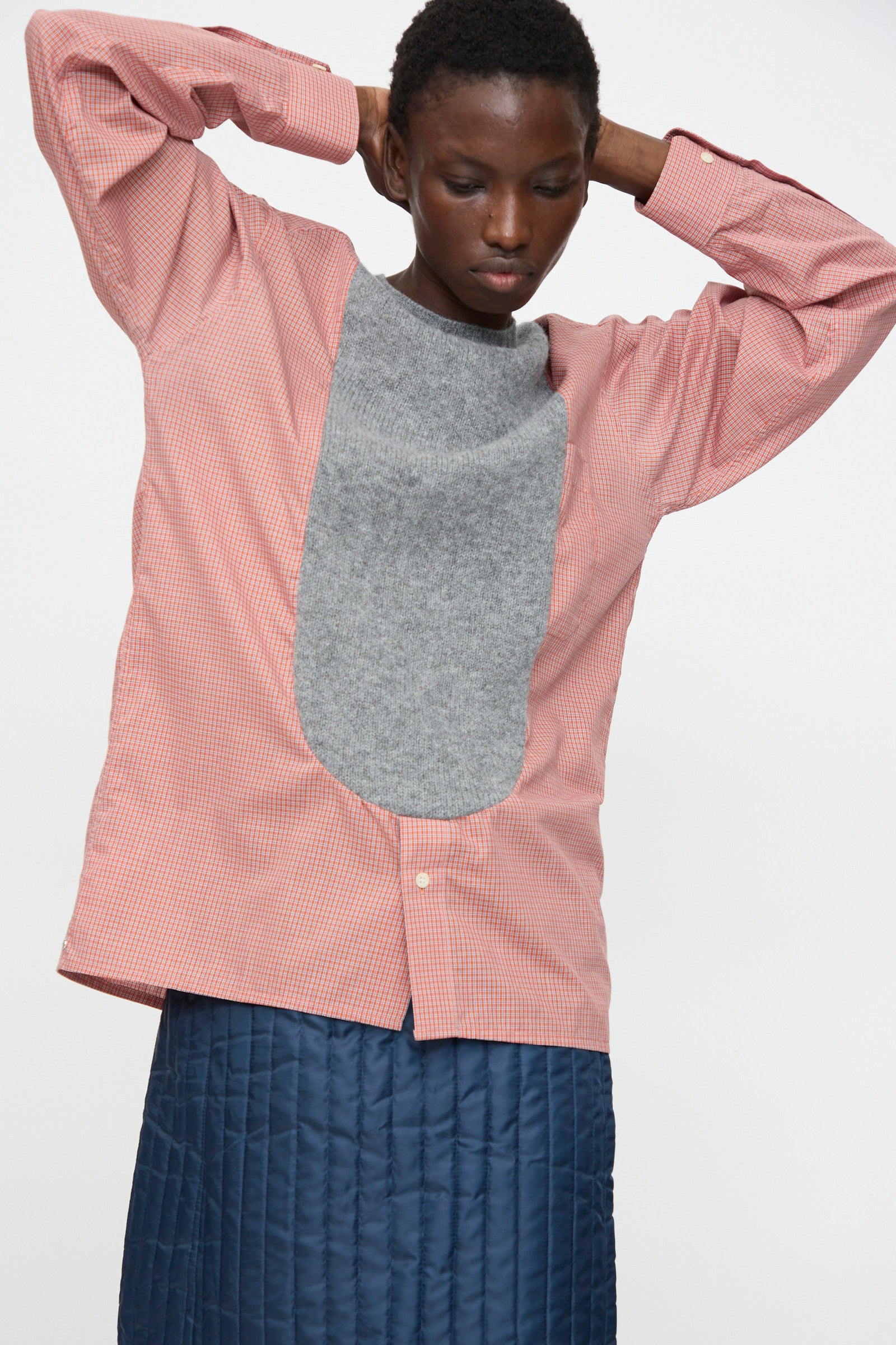 A person wears the Bless No. 68 Frontinsertpullover in Unique IV, featuring a red and white cotton poplin shirt with a large Shetland wool front panel, paired with a blue quilted skirt, standing against a plain background.