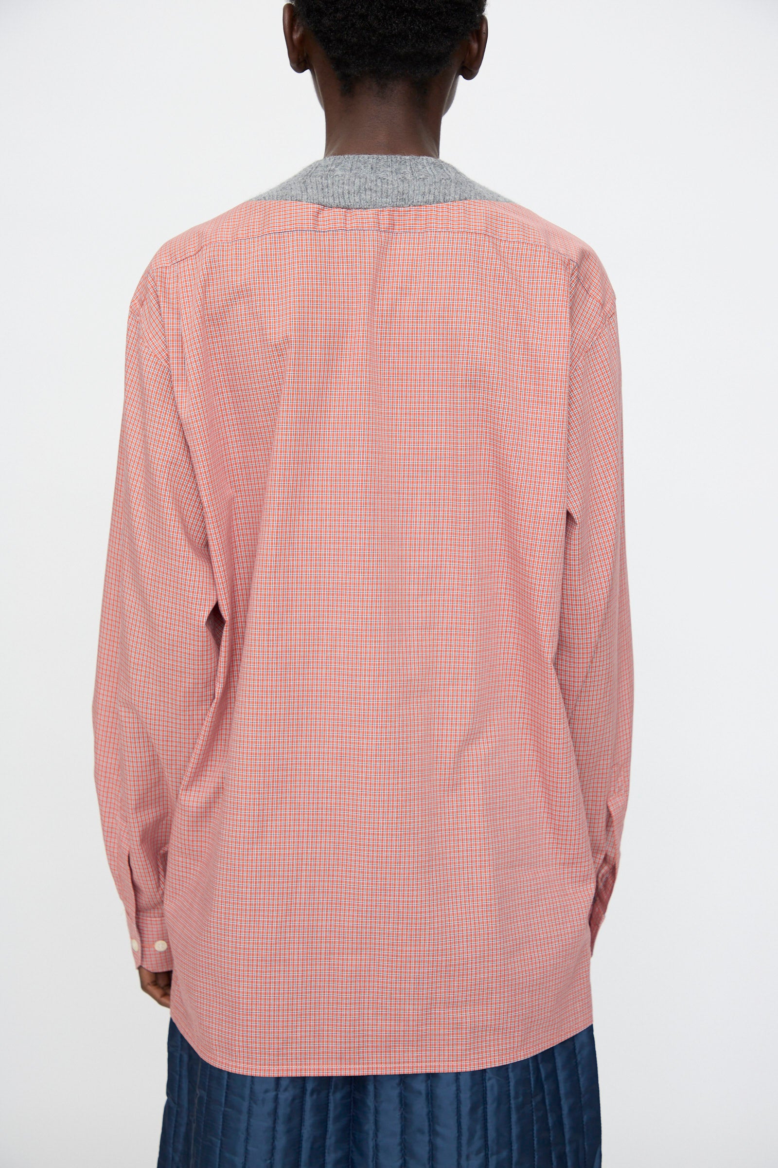 A person faces away, wearing Bless's No. 68 Frontinsertpullover in Unique IV—a red and white checkered cotton poplin shirt with a grey collar—paired with a blue quilted skirt, set against a plain white background.