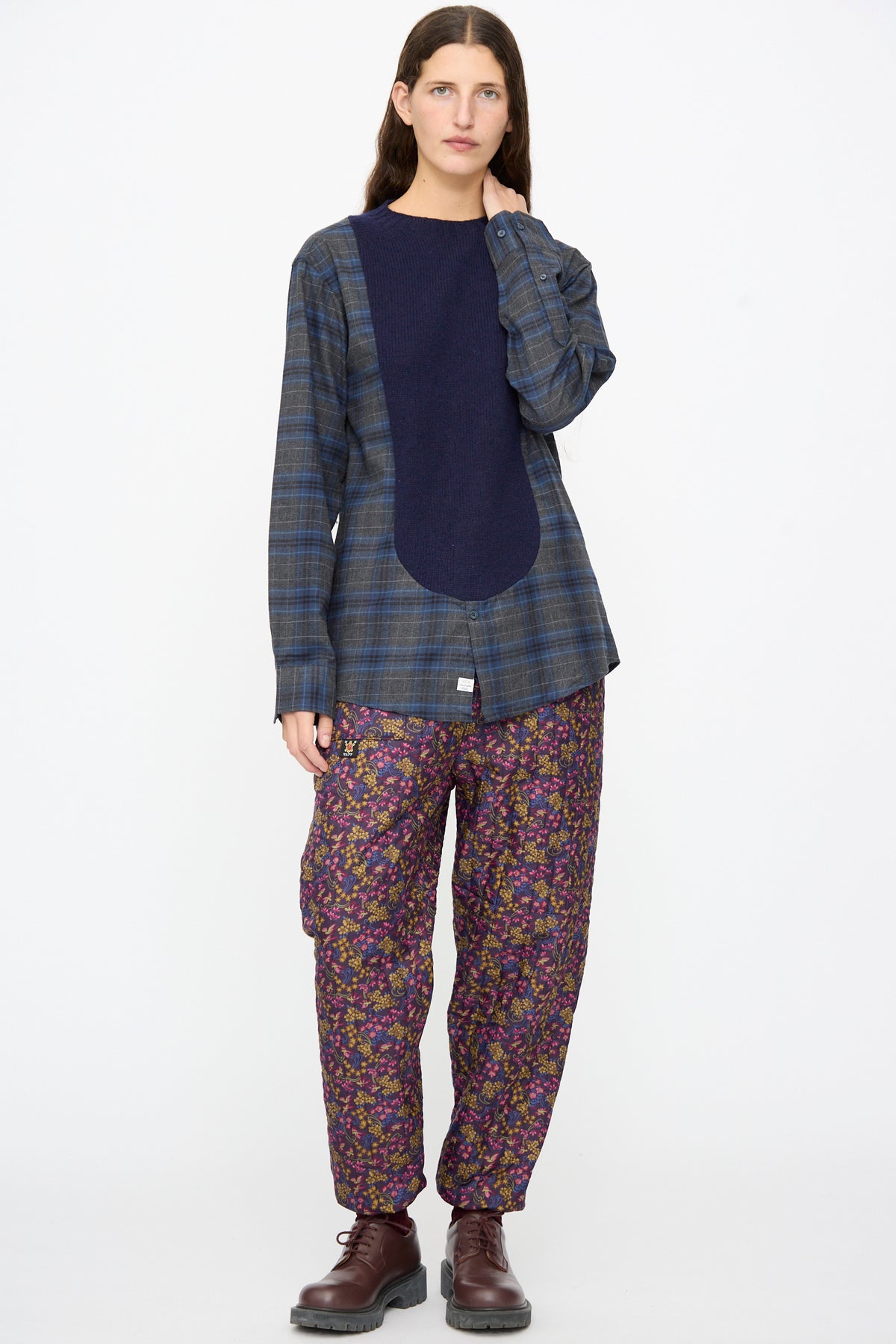 A model wearing a Bless No. 68 Frontinsertpullover in Unique IV over a blue and grey plaid flannel shirt, paired with floral print trousers and dark brown boots.
