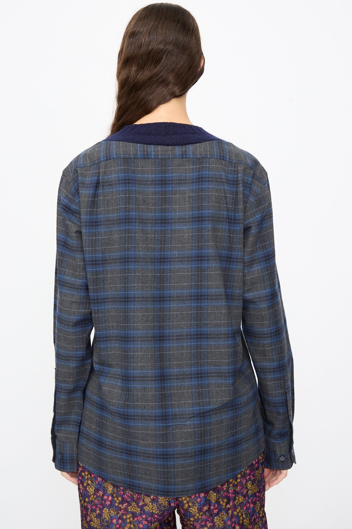 Back view of a person wearing a Bless No. 68 Frontinsertpullover in Unique IV, a blue and grey plaid flannel shirt with a ribbed dark blue collar, tucked into floral print pants.