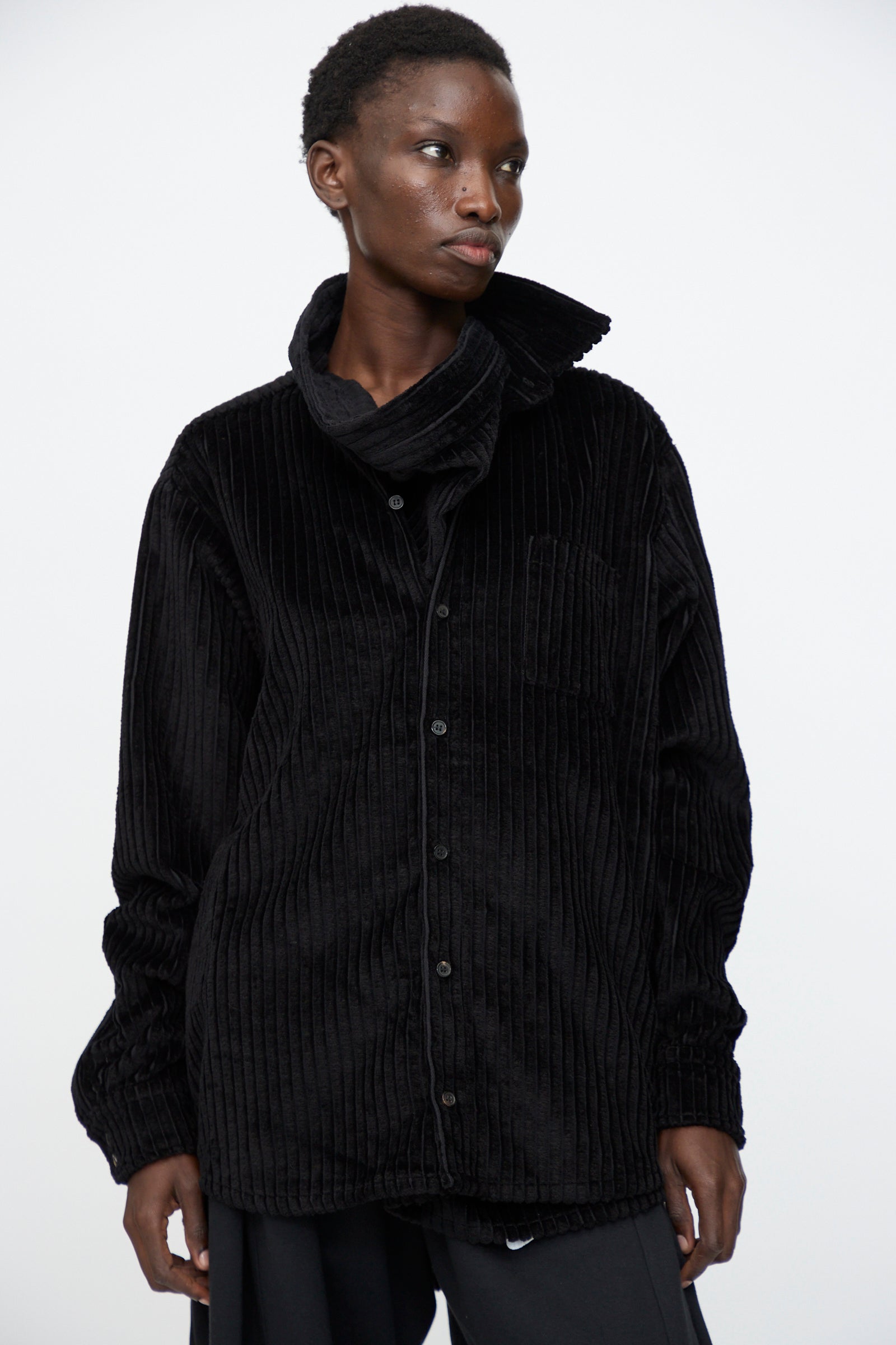 A person wears the Bless No. 68 Neckerchiefer in Black Corduroy—a high wrap-collar, button-up shirt—while standing against a plain white background.