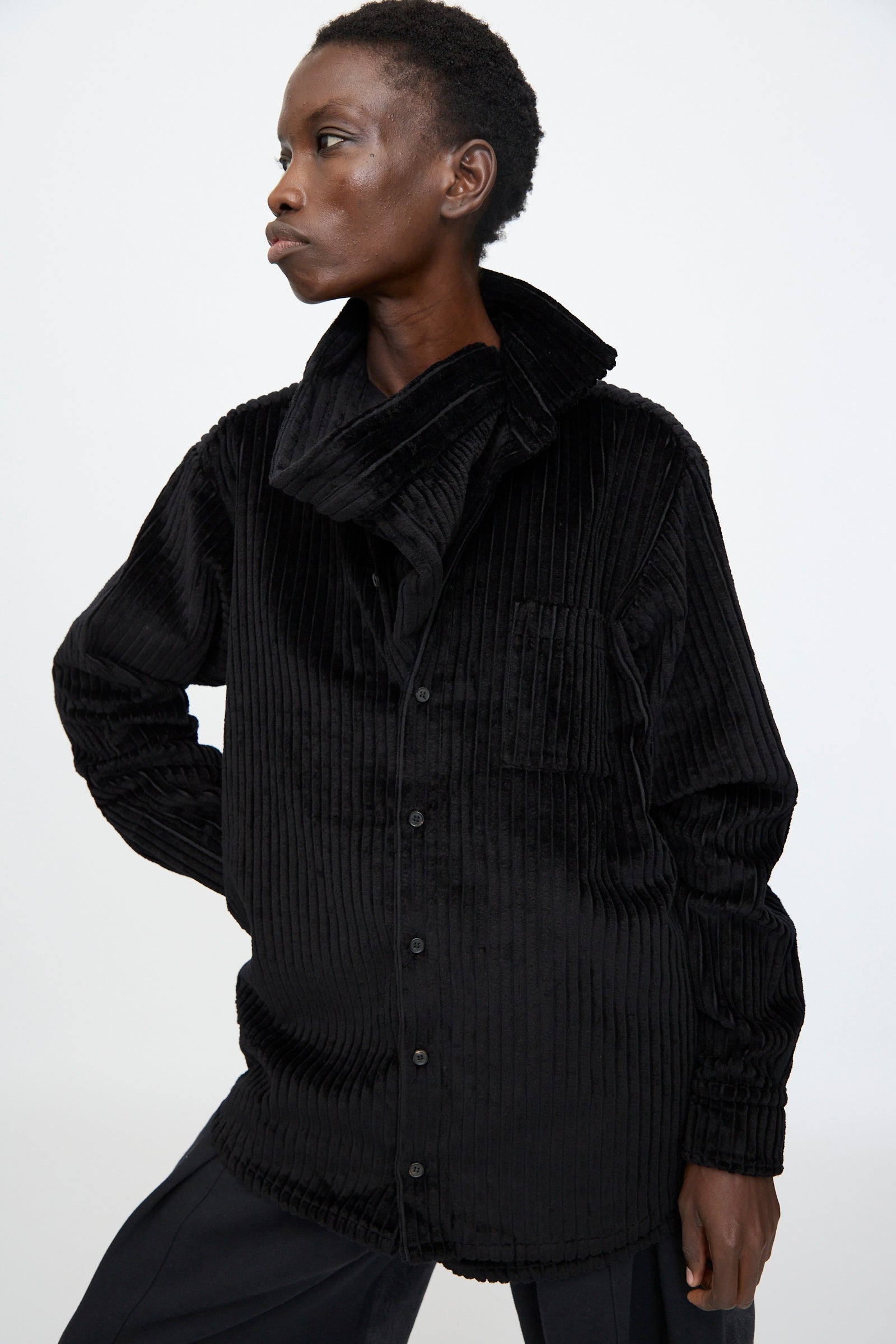 A model wears the Bless No. 68 Neckerchiefer in black corduroy, featuring a textured, button-up design and high collar, standing with one hand on their hip against a plain white background.
