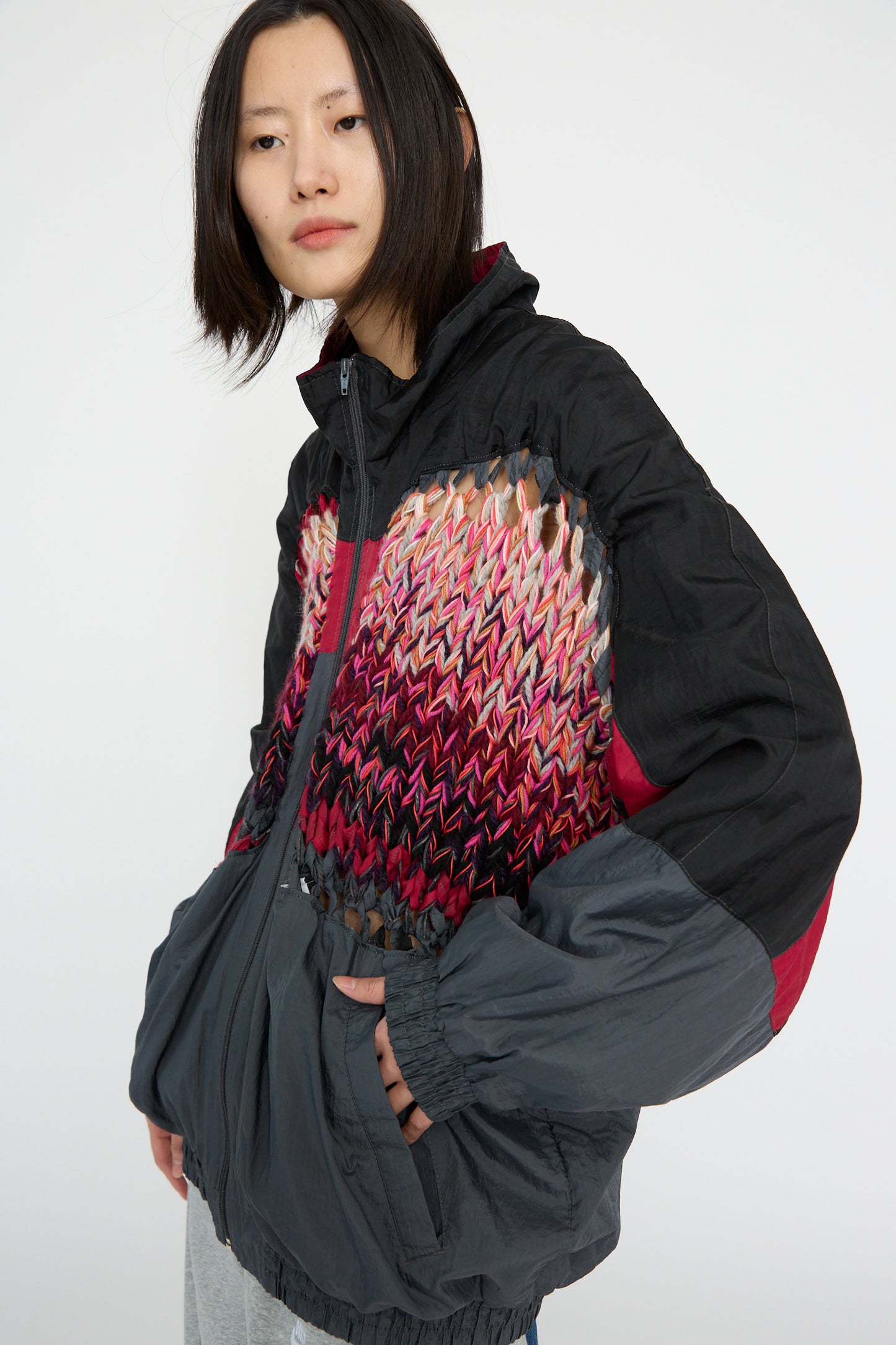 A person stands against a plain background wearing the Bless No. 70 Aknitter Unique Jacket in Multicolor Mix III,  with a hand in their pocket. 