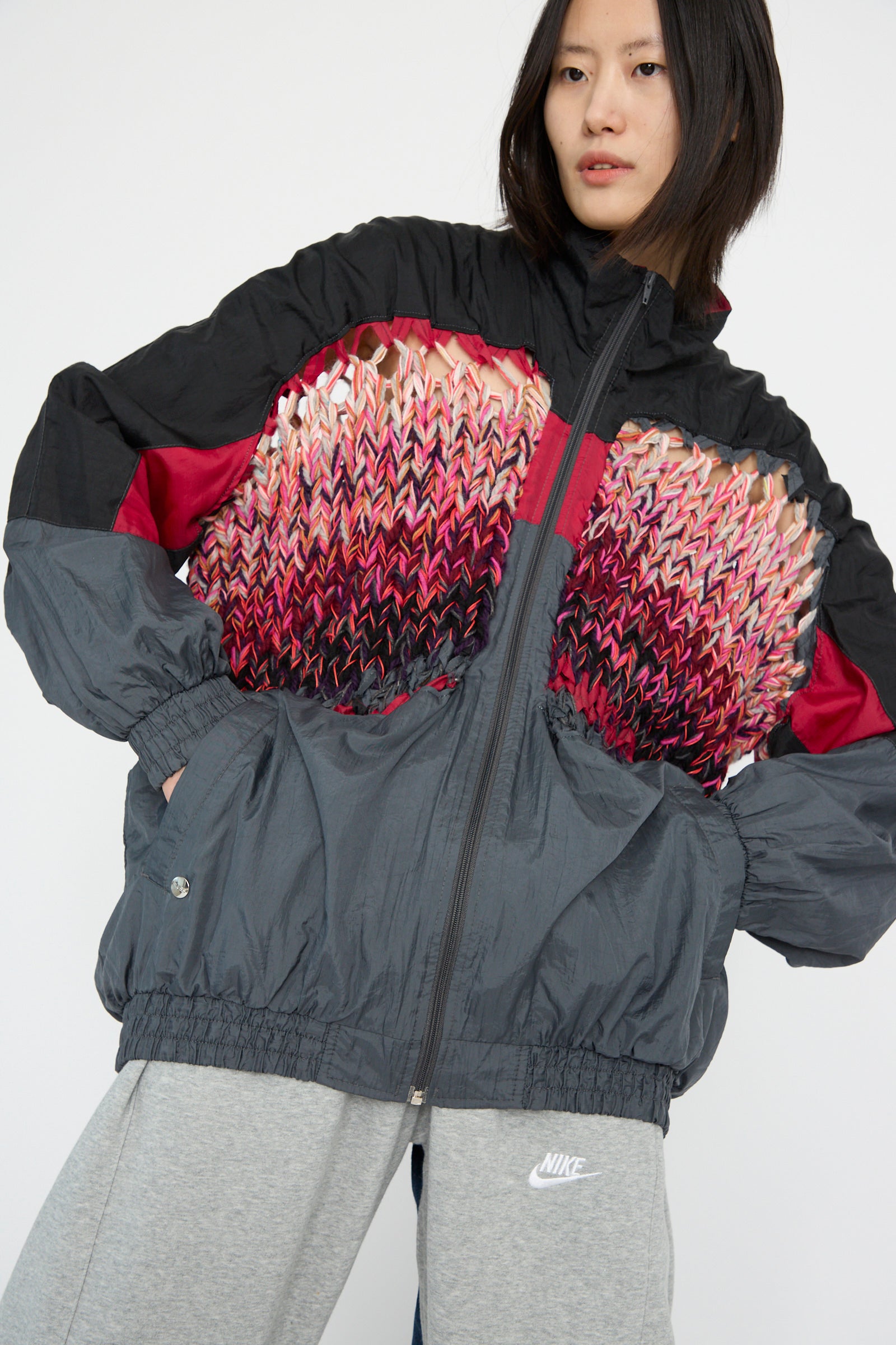 A model wears the No. 70 Aknitter Unique Jacket in Multicolor Mix III by Bless, showcasing knit patterns of red and pink alongside gray Nike sweatpants, against a plain backdrop.