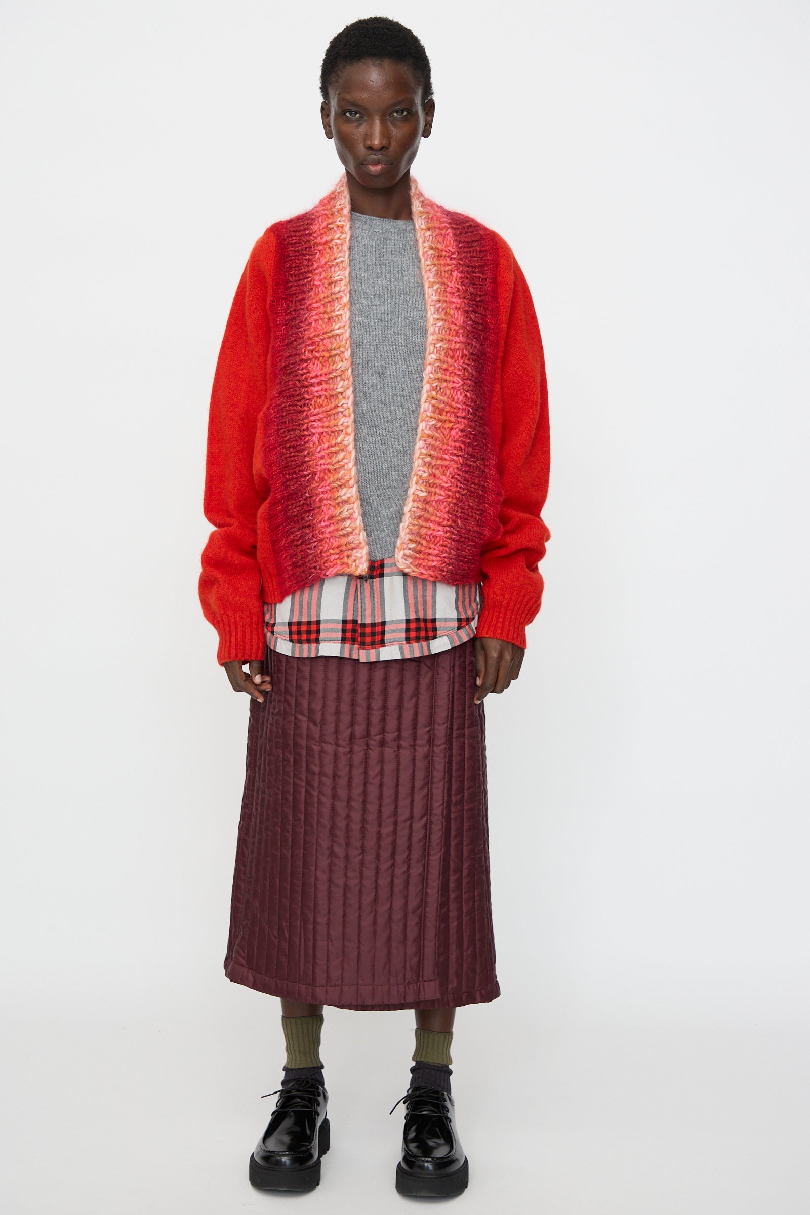 A person stands against a white background wearing Bless's No. 70 Passencardigan in Scarlet, layered over a gray sweater, plaid shirt, maroon quilted skirt, green socks, and black shoes.