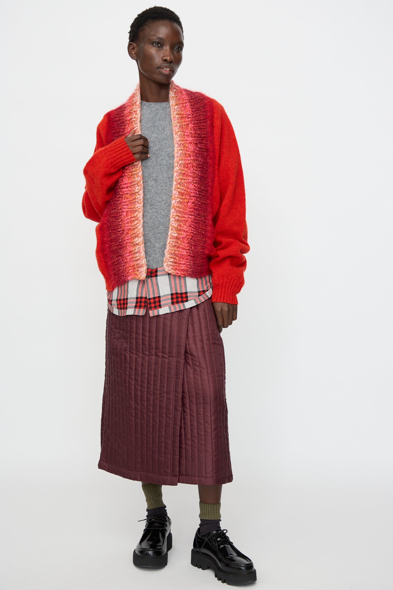A model wears the Bless No. 70 Passencardigan in Scarlet, paired with a gradient scarf, gray shirt, plaid layer, maroon quilted skirt, olive socks, and black shoes against a plain white background.