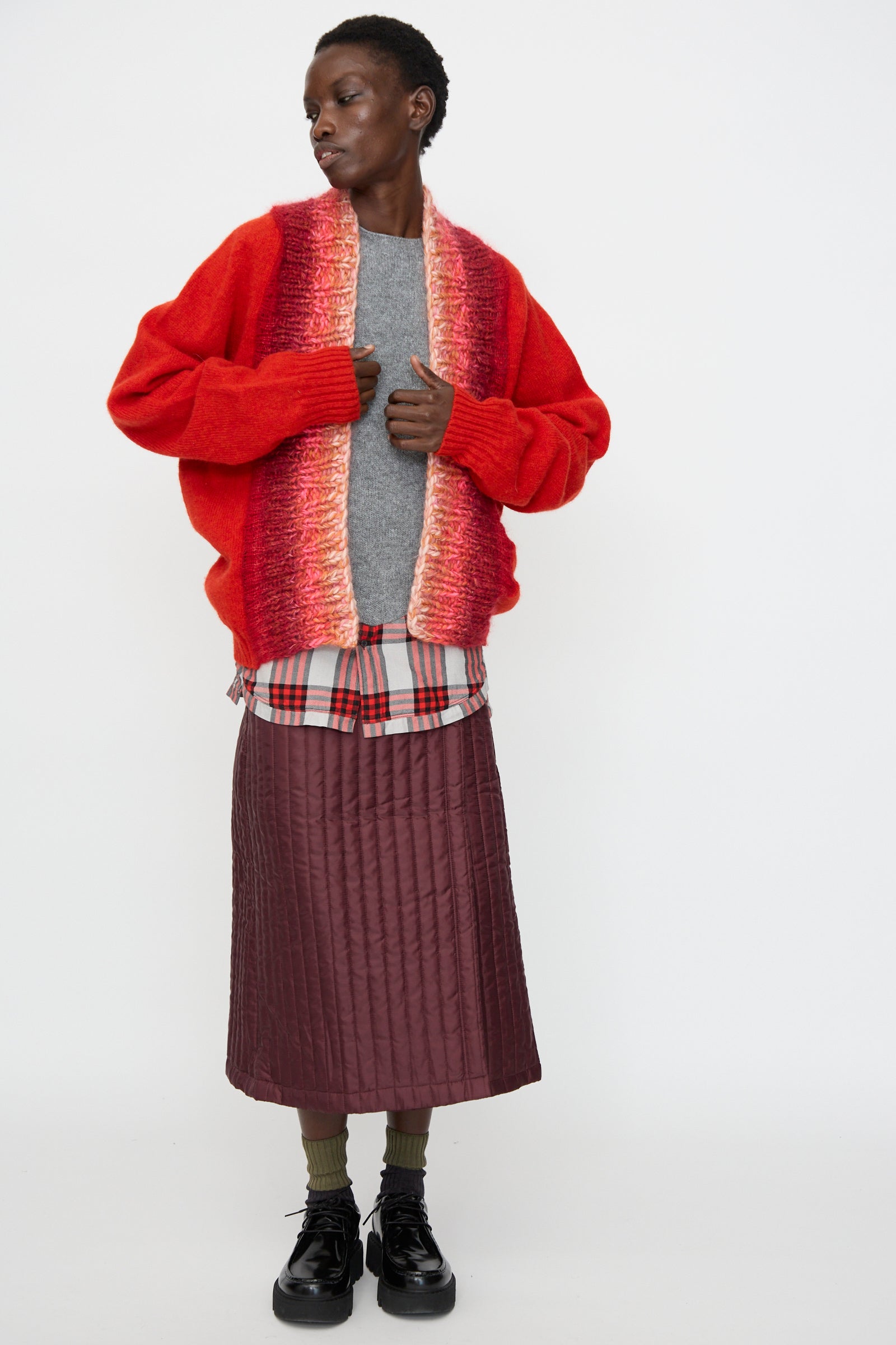 A person stands against a white background wearing the Bless No. 70 Passencardigan in Scarlet, a grey sweater, plaid shirt, maroon quilted skirt, green socks, and black shoes.