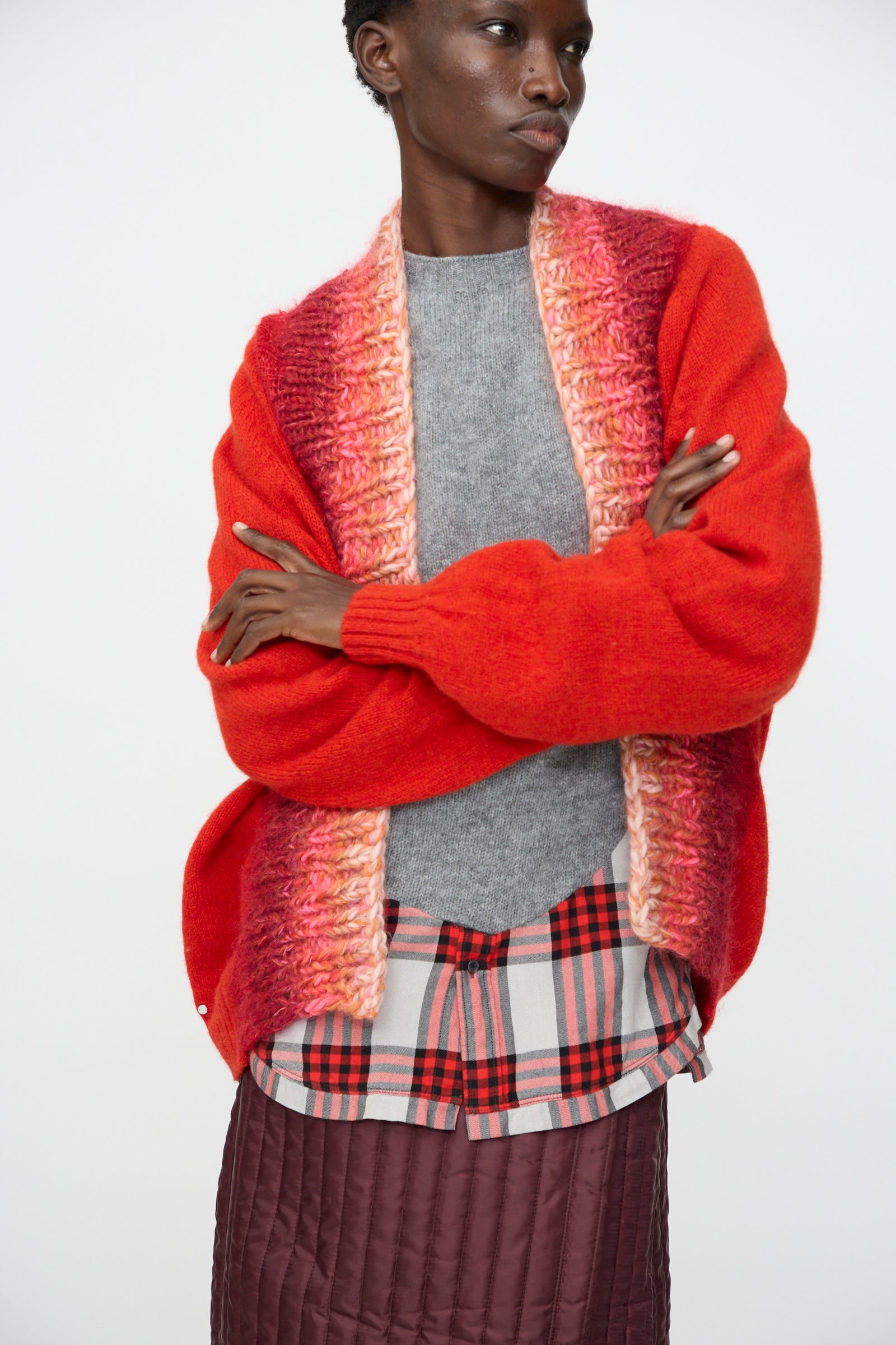 A person wears the Bless No. 70 Passencardigan in Scarlet—an open, hand-knitted red and pink mohair cardigan—layered over a gray sweater, plaid shirt, and burgundy quilted skirt, standing with arms crossed against a plain background.