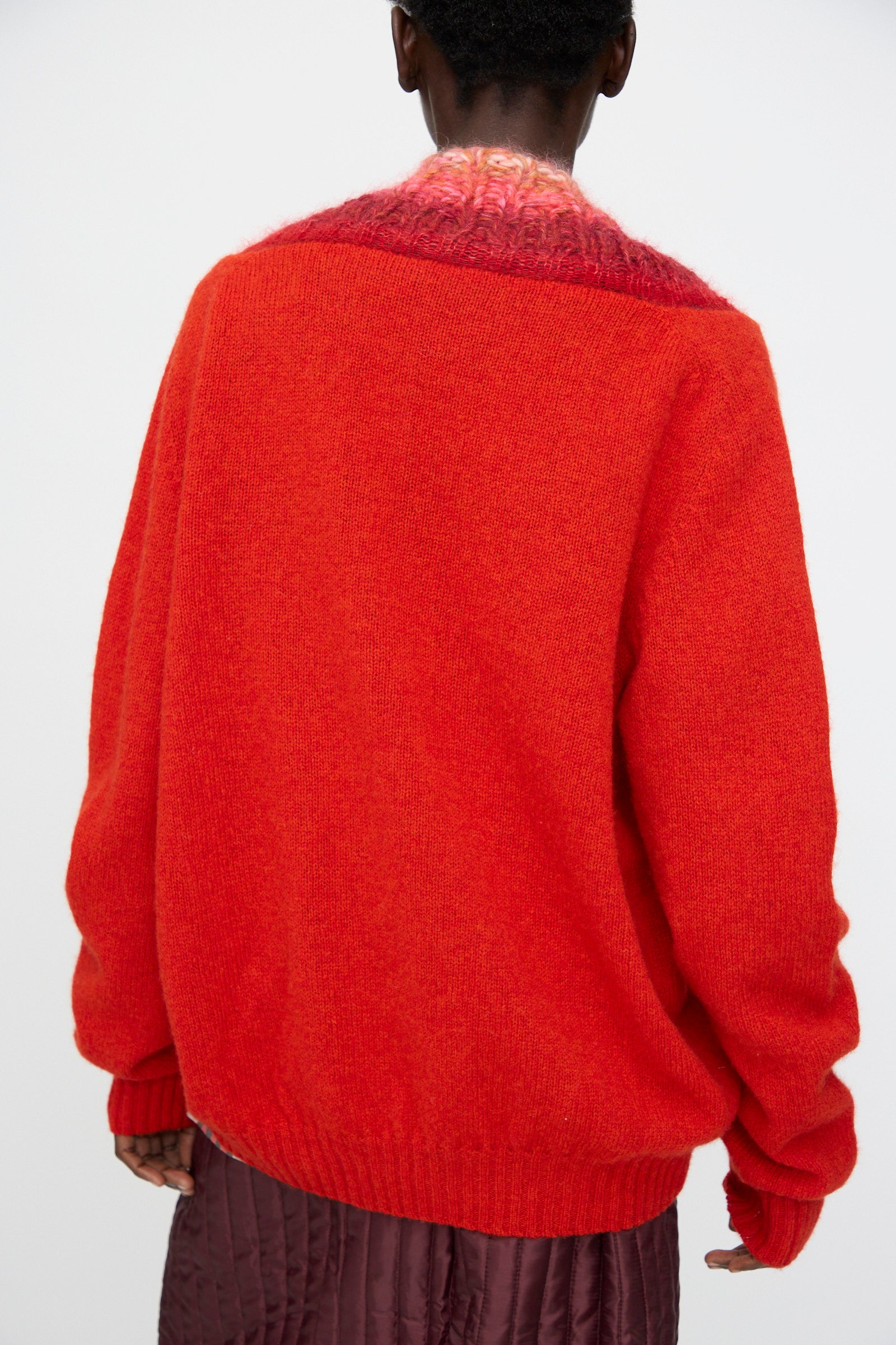 Model wears the Bless No. 70 Passencardigan in Scarlet—a loose-fitting red wool mohair sweater with gradient detailing at the shoulders—paired with a brown quilted skirt, shown from the back.