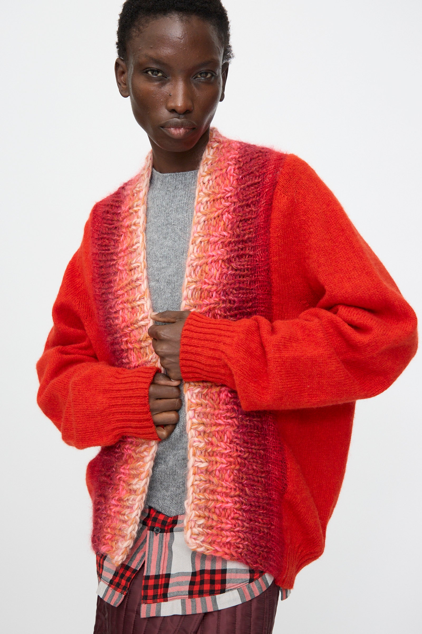 A person wears the Bless No. 70 Passencardigan in Scarlet, a hand-knitted wool mohair open cardigan, over a gray top and patterned skirt, standing against a plain background.