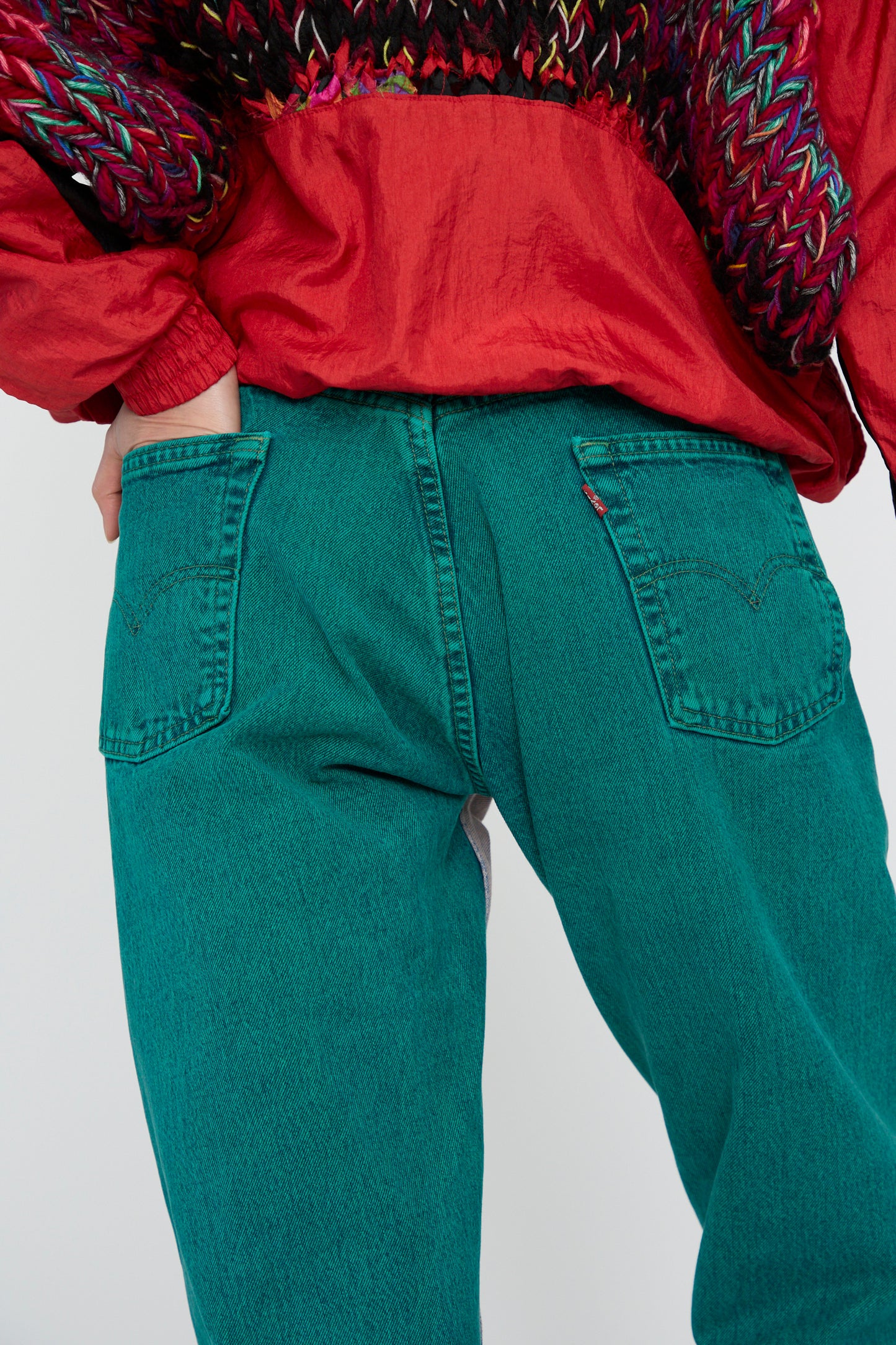 A close-up of a model wearing a red jacket and jeans from Bless's No. 73 Jeanspleatfront in Surprise I stands with hands in the back pockets against a plain background.