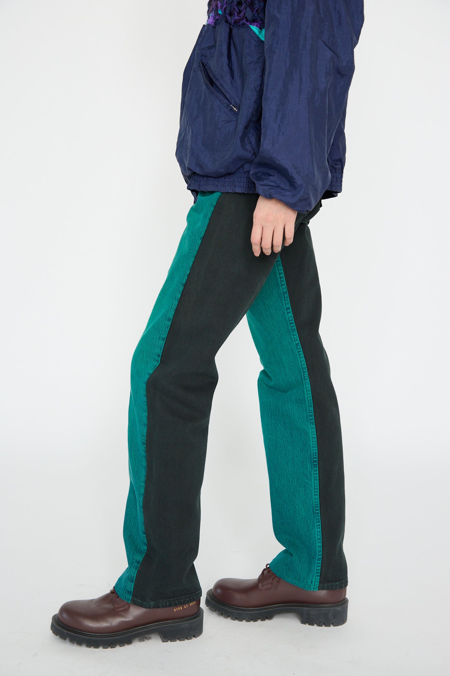 A model wears No. 73 Jeanspleatfront in Surprise II by Bless, teal and black pants, and brown shoes while standing sideways against a white background.