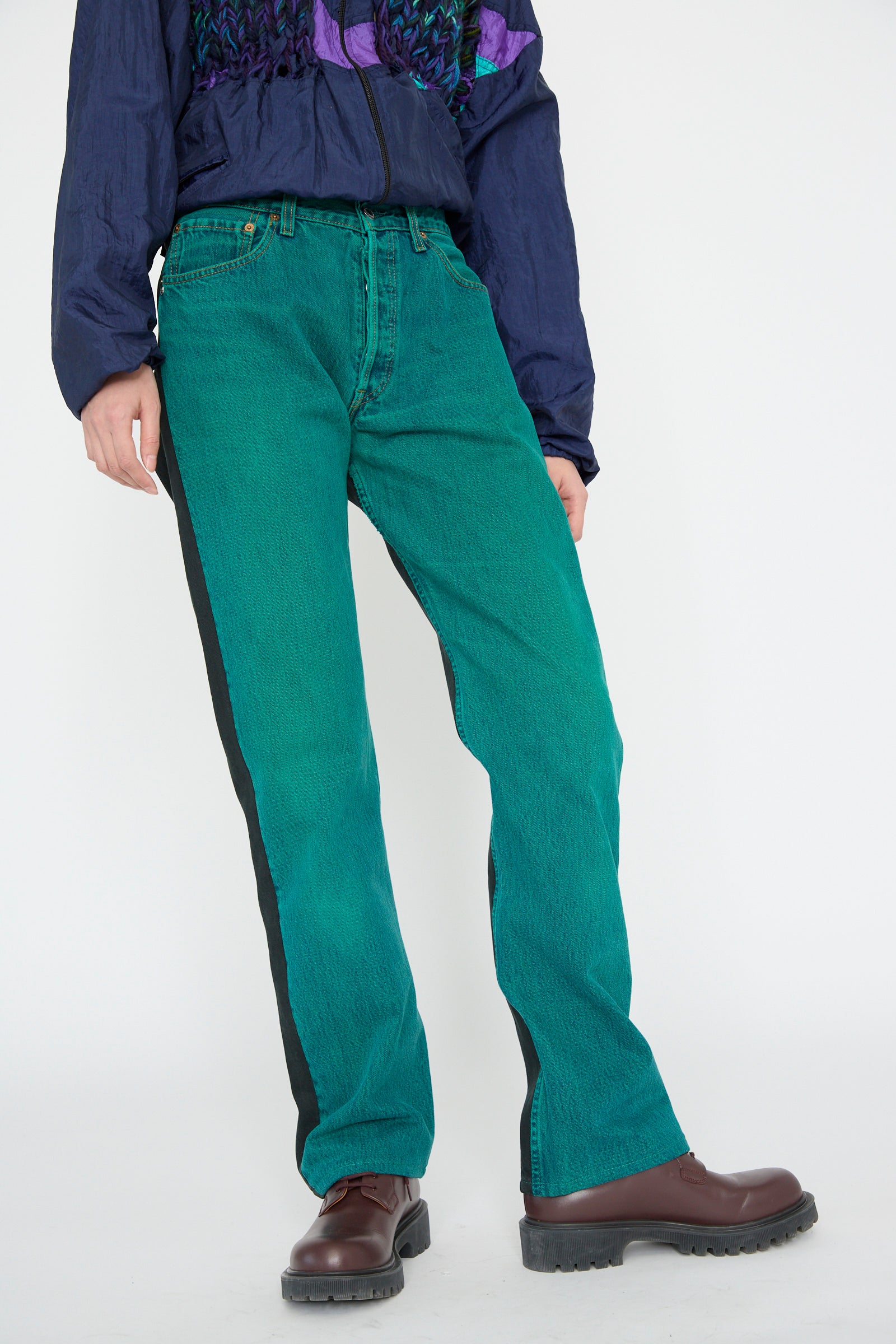 A model wearing Bless's No. 73 Jeanspleatfront in Surprise II, a relaxed-fit green pair with a  blue jacket and brown shoes stands against a plain background.