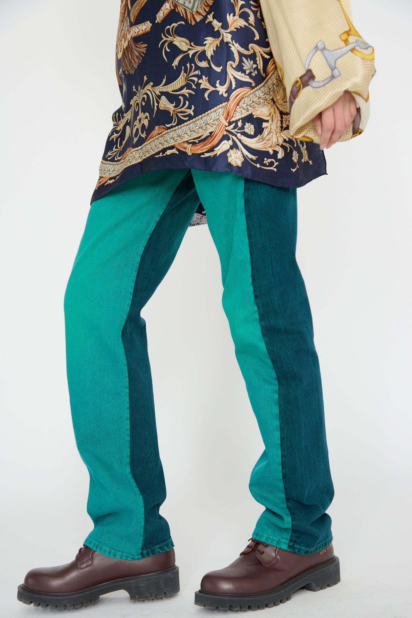 A model wearing the No. 73 Jeanspleatfront in Surprise III by Bless, featuring teal and green color-block up-cycled vintage denim, an ornate patterned top, and brown shoes, walking against a plain background.