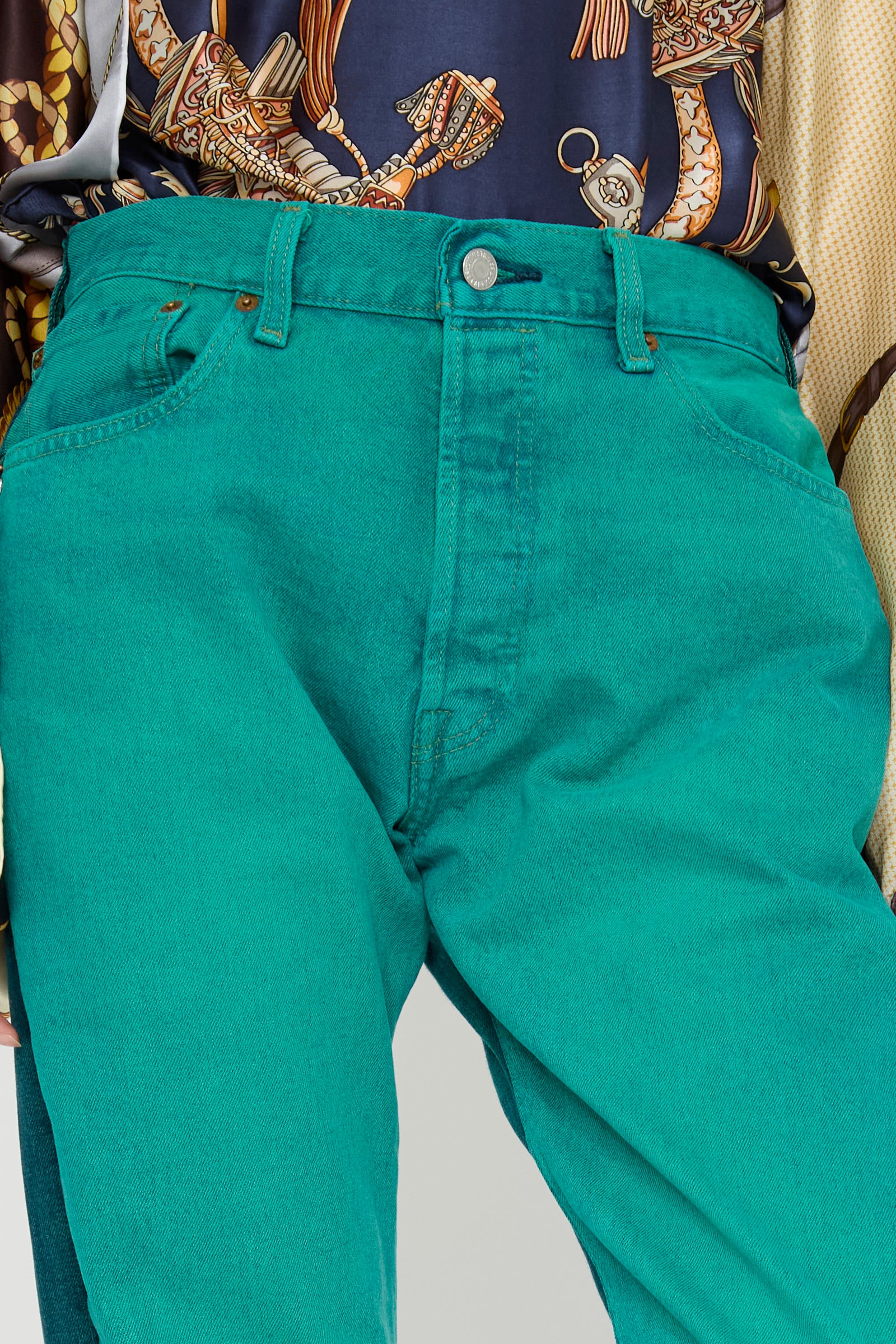 A close-up of a person wearing turquoise, straight-leg jeans and a patterned shirt with ropes and chains. The jeans are the No. 73 Jeanspleatfront in Surprise III by Bless, made from up-cycled vintage.