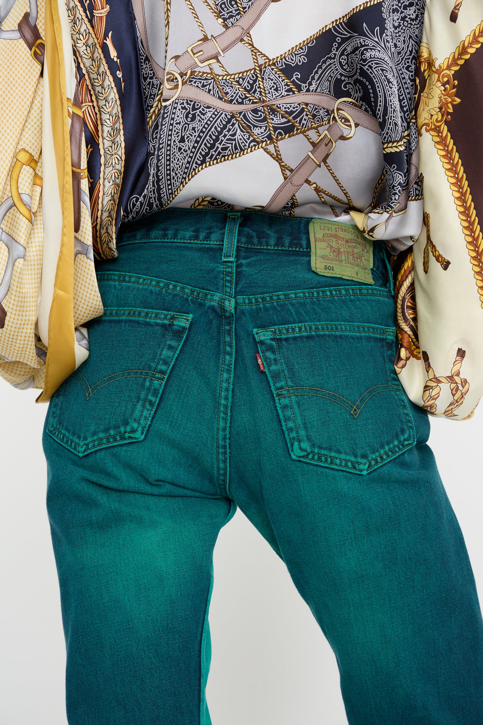 A person wearing Bless No. 73 Jeanspleatfront in Surprise III stands with their back to the camera, styled in straight-leg teal jeans and a patterned shirt featuring a chains and ropes design, all made in France.