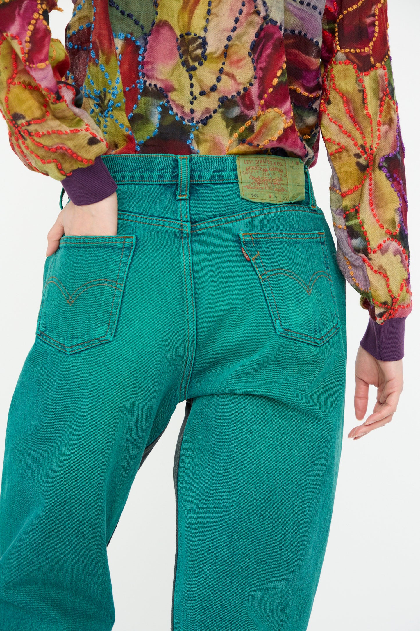 A model is wearing Bless's No. 73 Jeanspleatfront in Surprise IV denim jeans, featuring a teal vintage up-cycled straight leg and relaxed fit, paired with a colorful patterned shirt, viewed from behind.