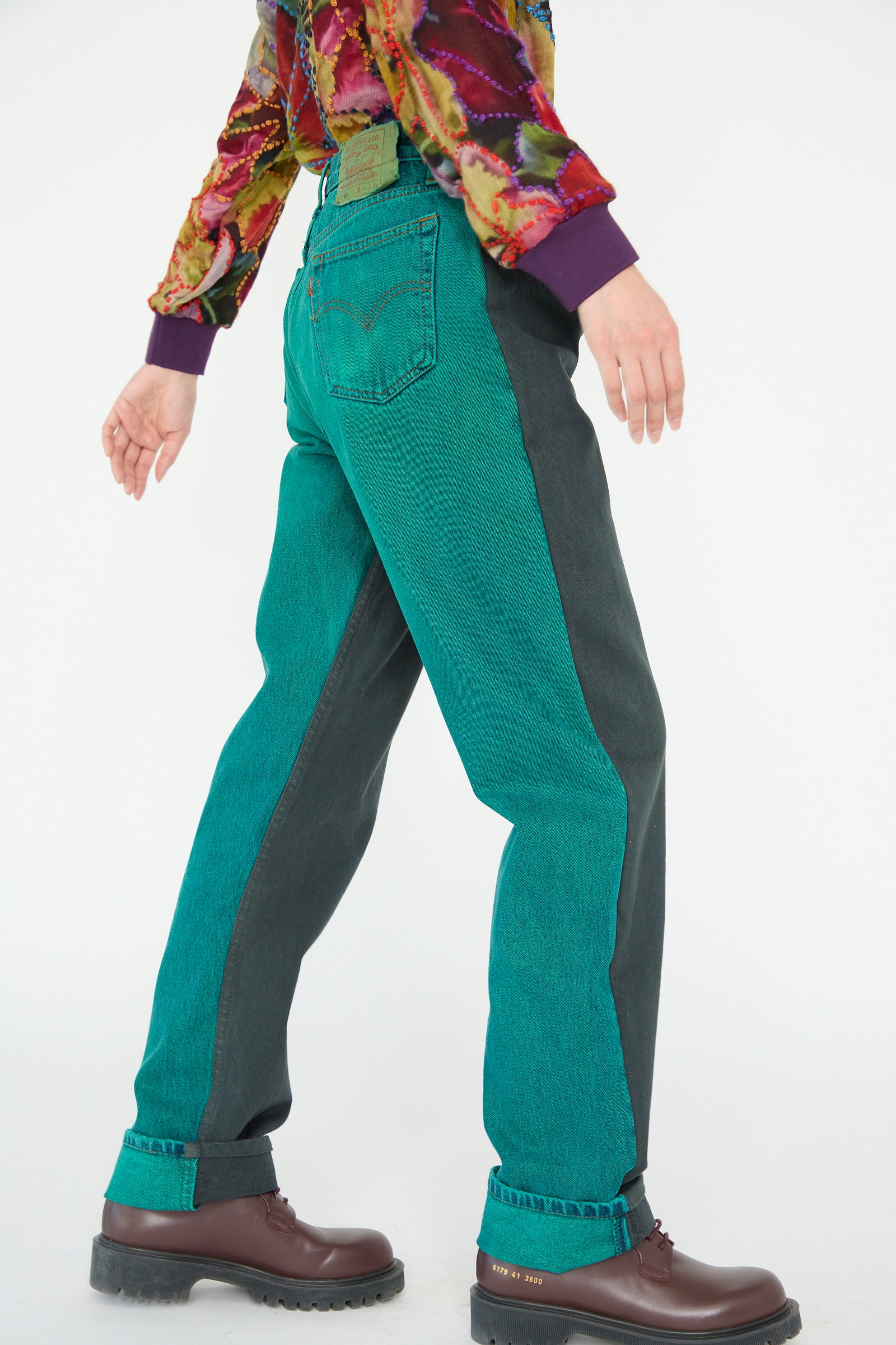 A model wearing Bless's No. 73 Jeanspleatfront in Surprise IV and a colorful top stands against a white background, walking to the side.