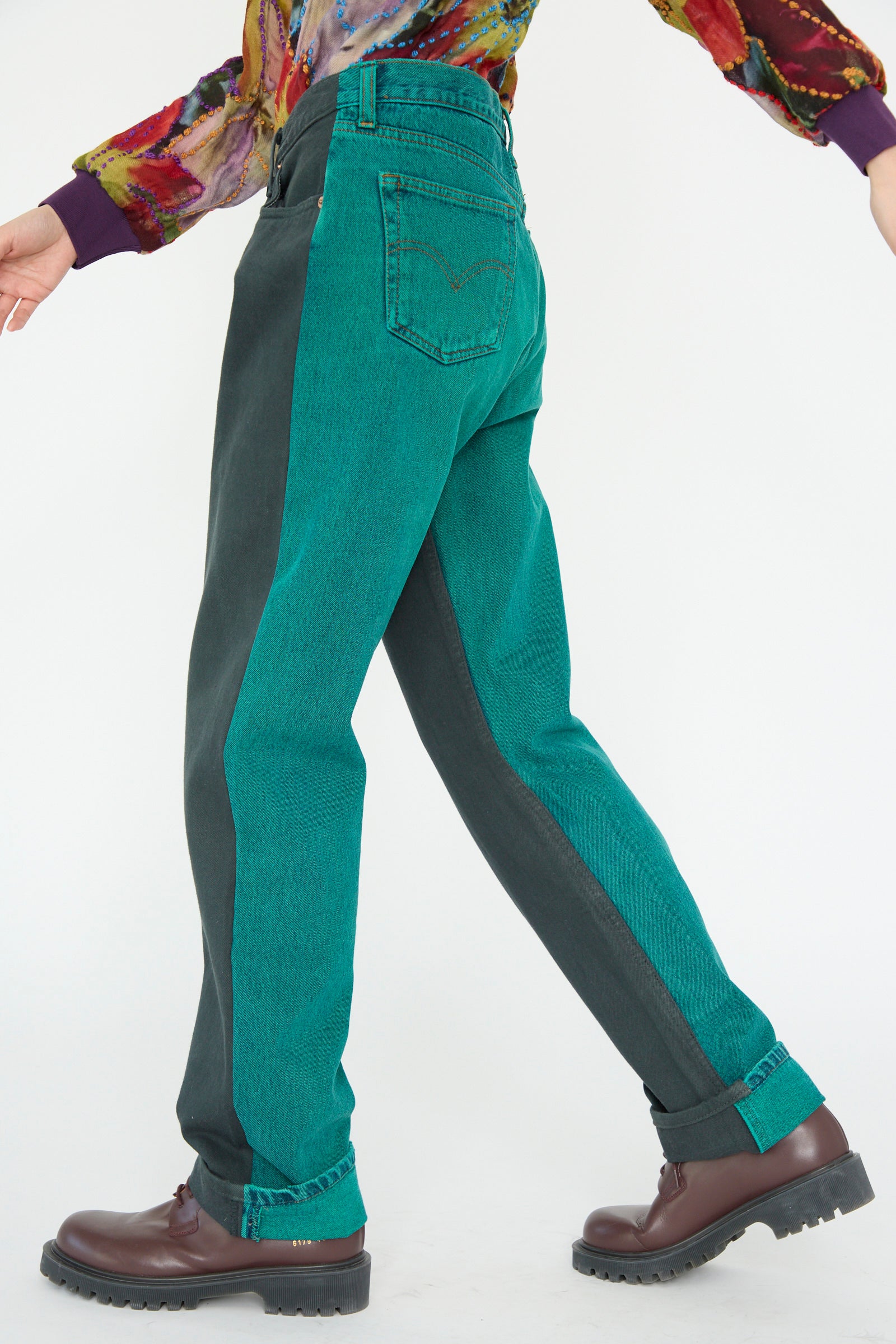 A person wearing Bless's No. 73 Jeanspleatfront in Surprise IV, a two-toned teal and dark green up-cycled vintage denim with a relaxed fit, paired with a colorful shirt and brown leather shoes, walks to the left.