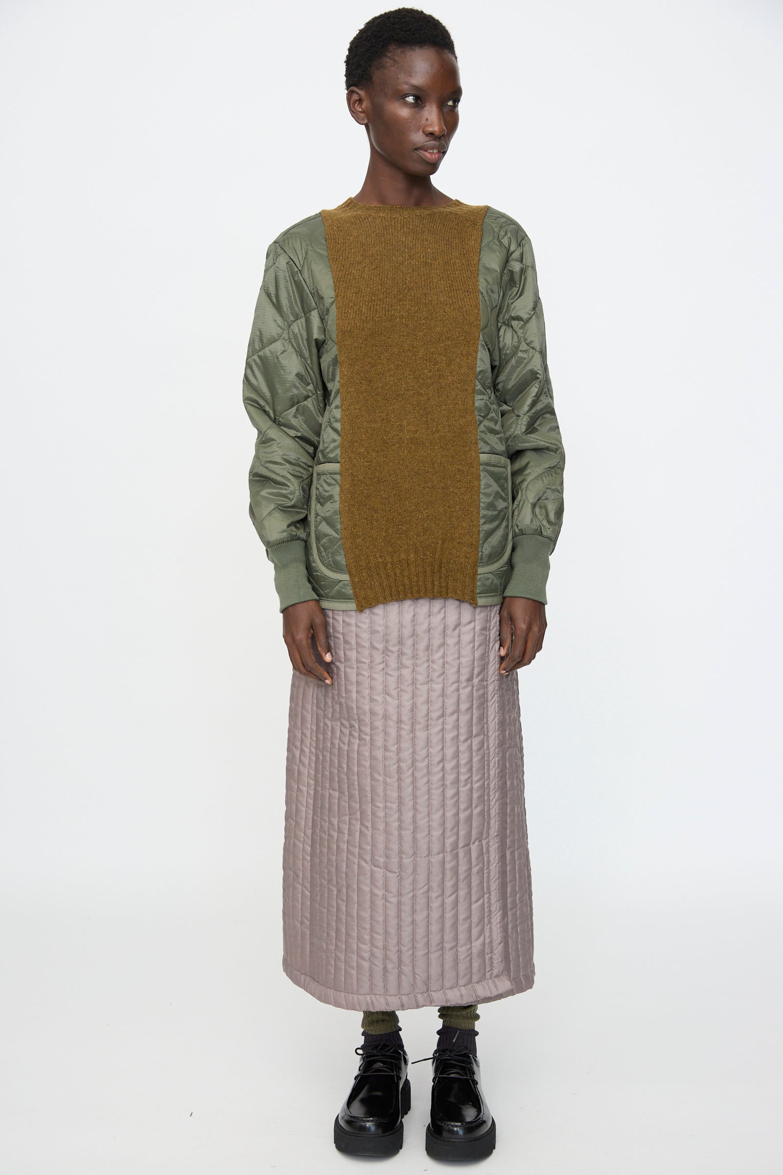 A person wears the Bless No. 78 Liningknitter Sweater in Olive, a long light pink quilted skirt, and black shoes while standing against a plain white background.