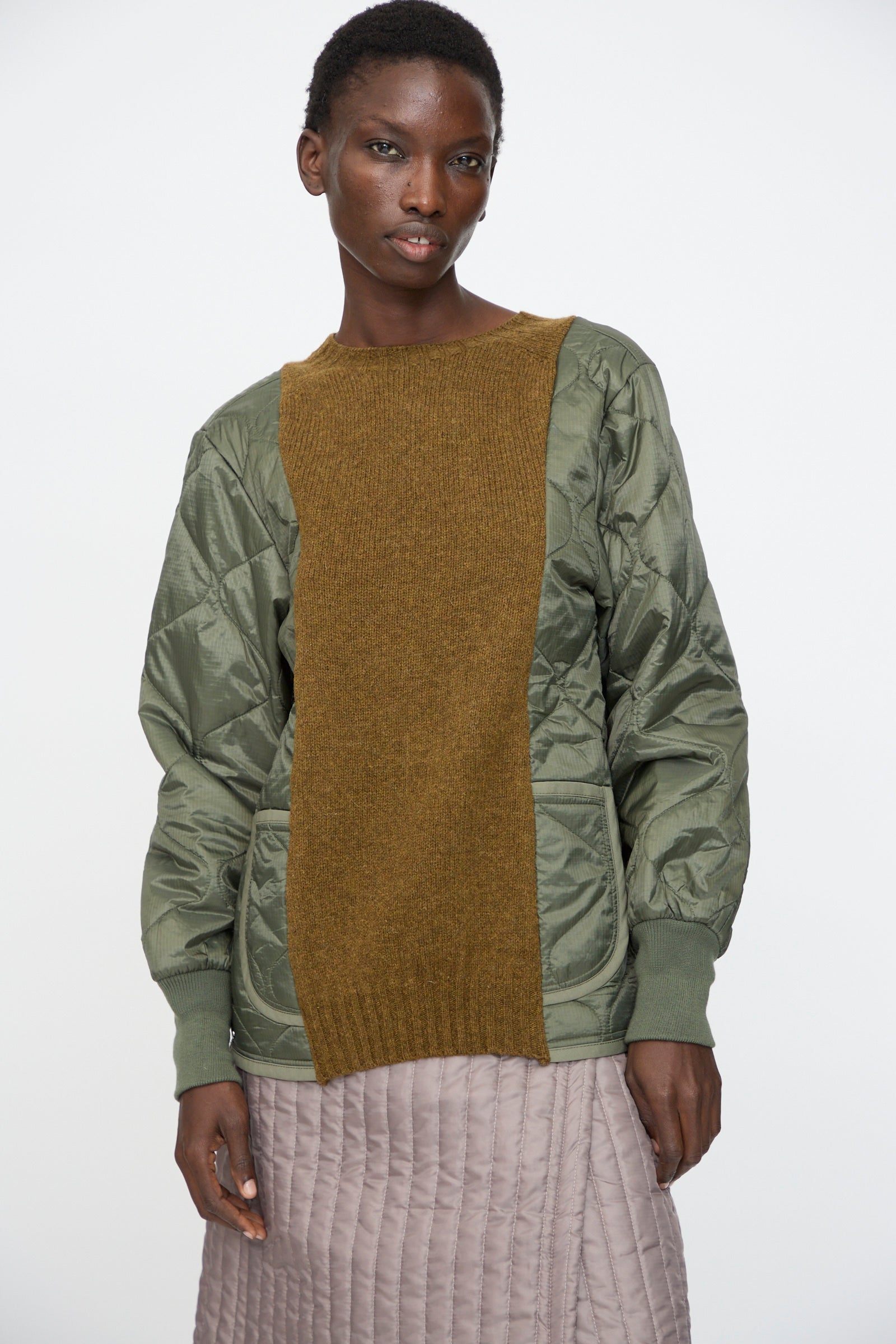 A person stands against a plain white background wearing Bless's No. 78 Liningknitter Sweater in Olive, styled under a green quilted jacket with a brown knit panel and paired with a light purple quilted skirt.