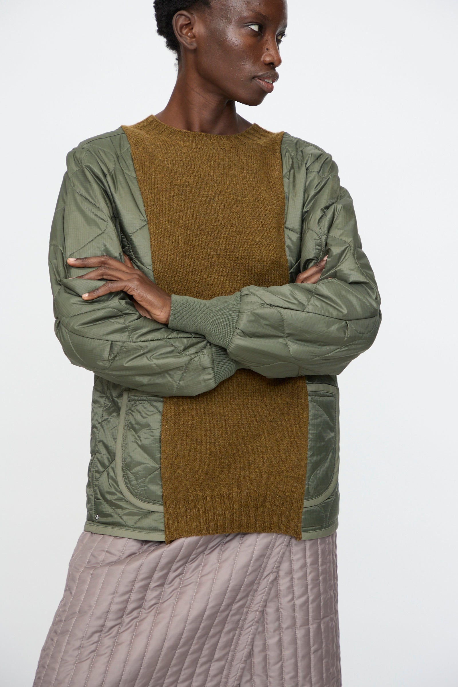 A person in the Bless No. 78 Liningknitter Sweater in Olive, layered under a green quilted jacket with brown knit panels and paired with a light brown quilted skirt, stands arms crossed against a plain background.